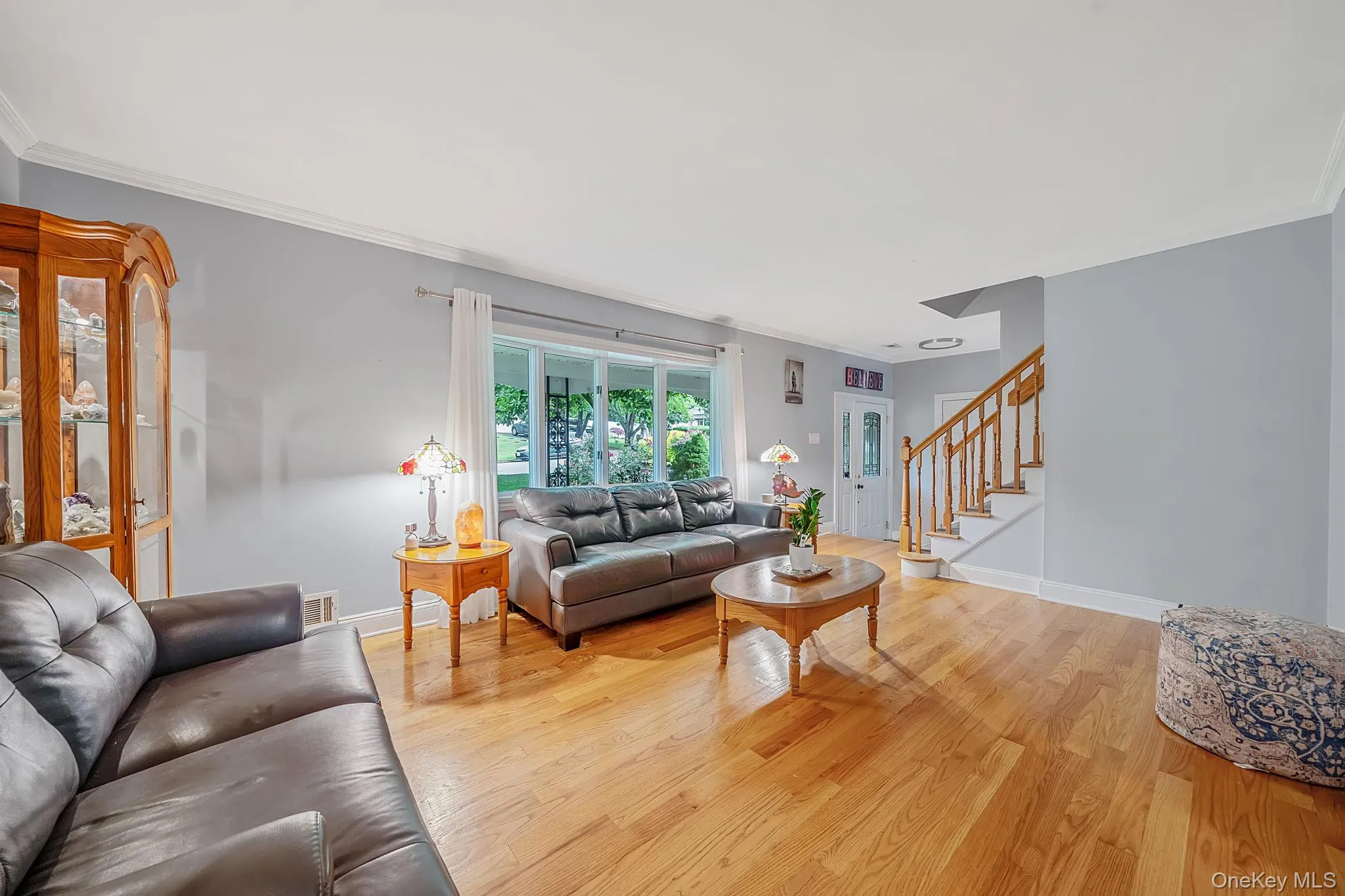 Spacious living area featuring hardwood flooring, a prominent staircase with wood railings, a large picture window, and crown molding Spacious living area featuring hardwood flooring, a prominent staircase with wood railings, a large picture window, and crown molding
