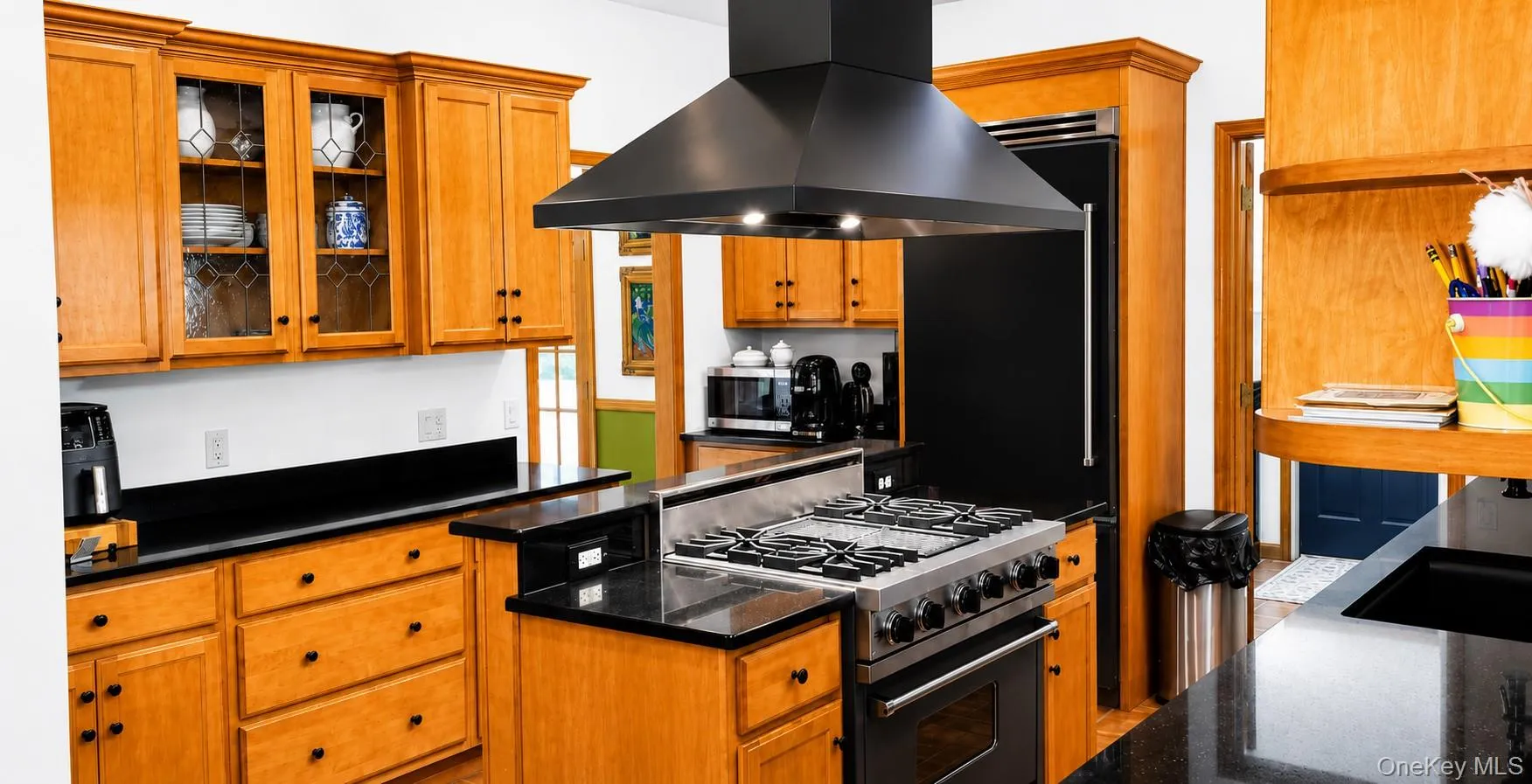 Gourmet kitchen featuring a stainless steel gas range with a black hood vent, black countertops, wood cabinetry, and a built-in black refrigerator Gourmet kitchen featuring a stainless steel gas range with a black hood vent, black countertops, wood cabinetry, and a built-in black refrigerator