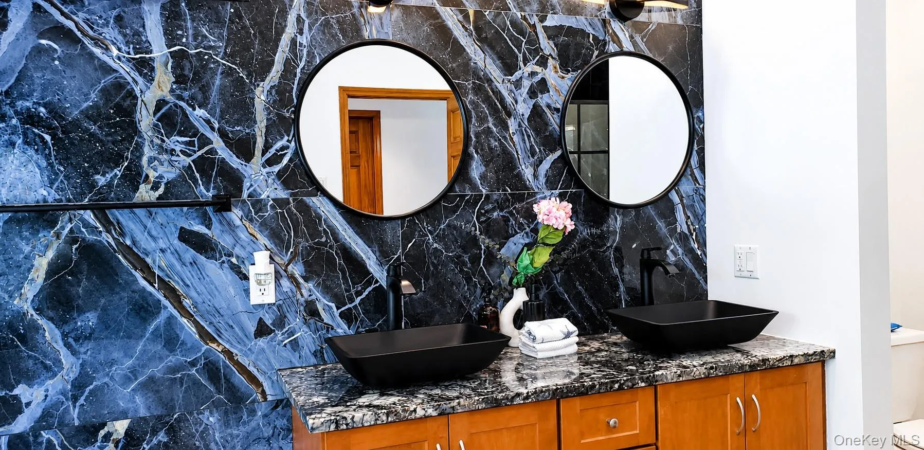 Double vanity featuring a black and blue veined accent wall, two vessel sinks, and wood-finish cabinetry Double vanity featuring a black and blue veined accent wall, two vessel sinks, and wood-finish cabinetry