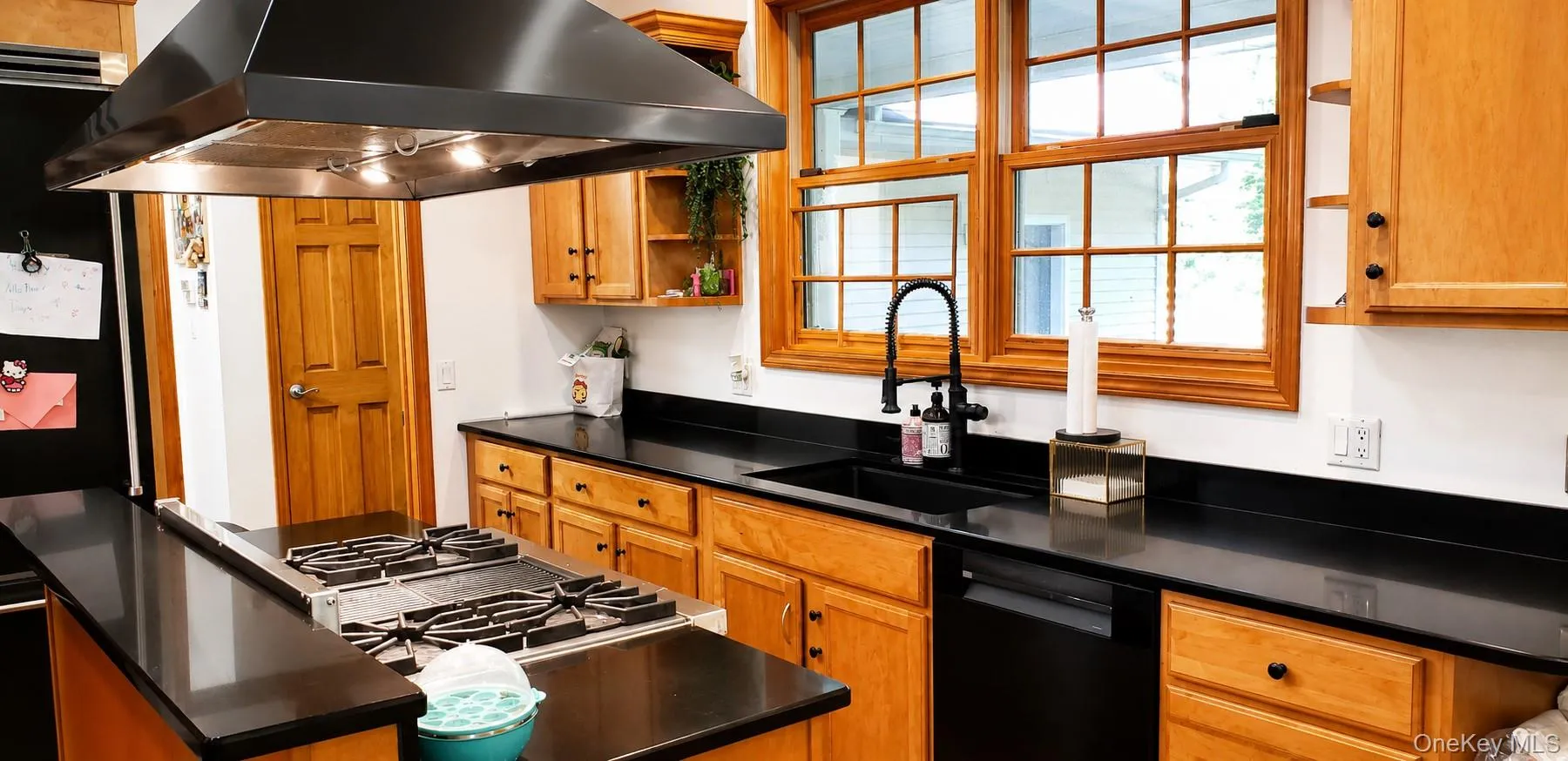 Kitchen featuring a built-in gas range with an overhead exhaust hood, black countertops, wood-finish cabinetry, a black sink with a matte black faucet, and multiple windows Kitchen featuring a built-in gas range with an overhead exhaust hood, black countertops, wood-finish cabinetry, a black sink with a matte black faucet, and multiple windows