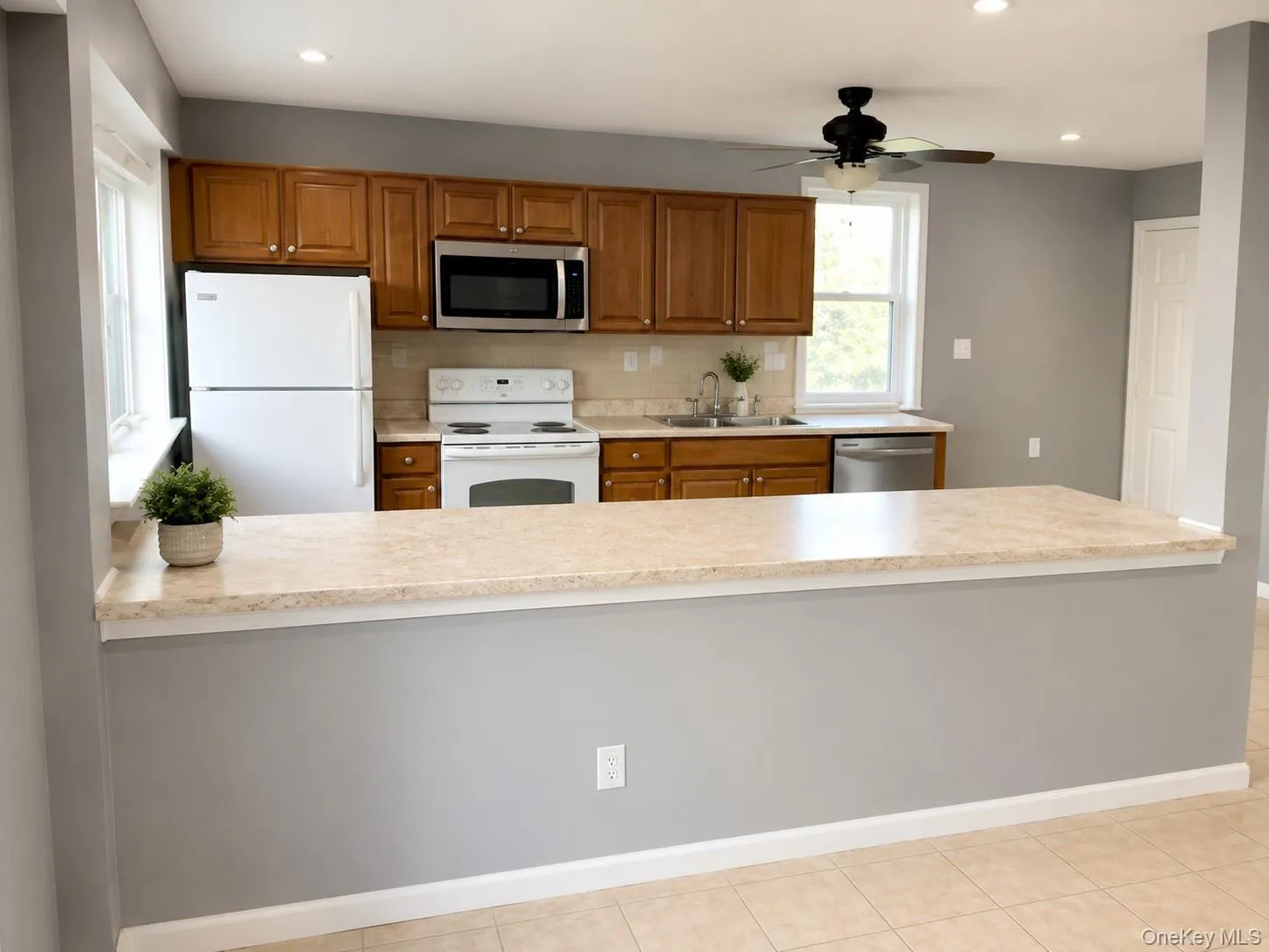 Kitchen featuring wood cabinetry, white appliances, stainless steel microwave and dishwasher, and a peninsula with a light-toned countertop Kitchen featuring wood cabinetry, white appliances, stainless steel microwave and dishwasher, and a peninsula with a light-toned countertop