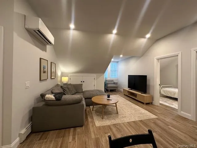 Living room featuring wood finished floors, a baseboard heating unit, a wall mounted AC, lofted ceiling, and recessed lighting Living room featuring wood finished floors, a baseboard heating unit, a wall mounted AC, lofted ceiling, and recessed lighting