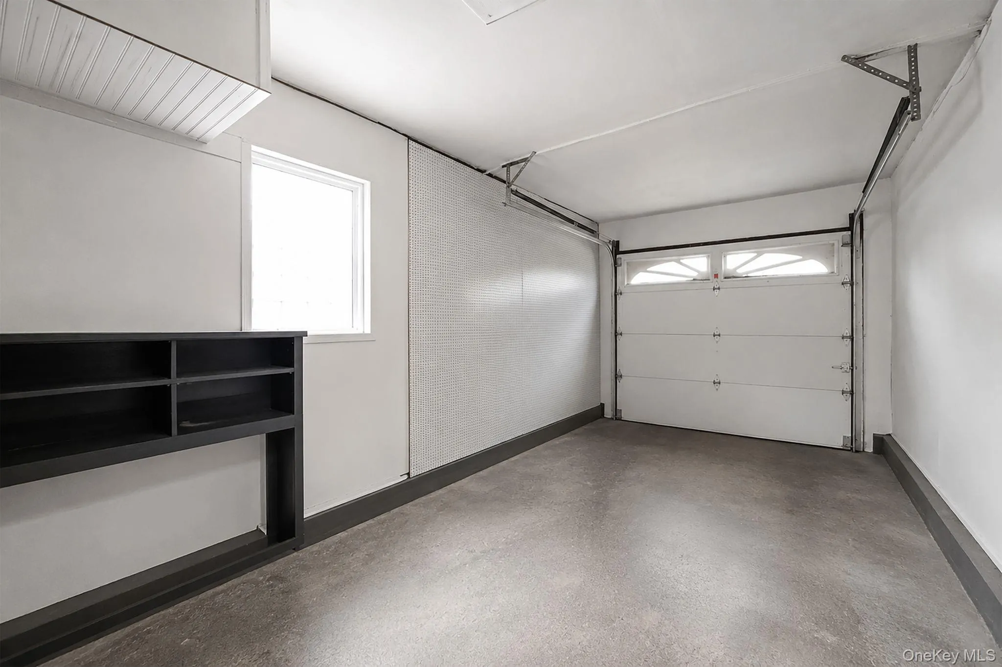 Attached one-car garage featuring a concrete floor, built-in shelving, an exterior window, and an overhead garage door with decorative lites Attached one-car garage featuring a concrete floor, built-in shelving, an exterior window, and an overhead garage door with decorative lites