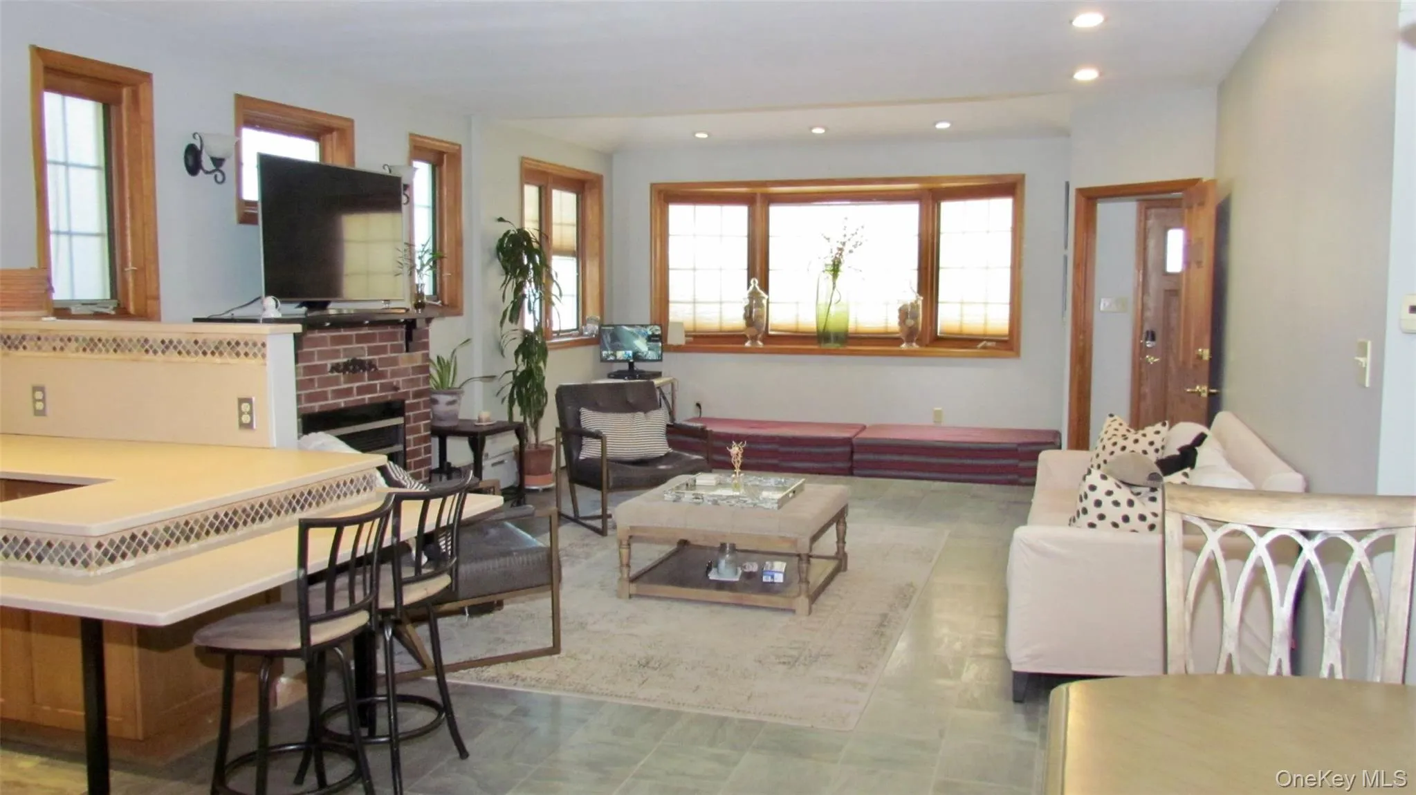 Open concept living area featuring tile flooring, a brick fireplace, built-in breakfast bar with patterned trim, wood-framed windows, and recessed lighting Open concept living area featuring tile flooring, a brick fireplace, built-in breakfast bar with patterned trim, wood-framed windows, and recessed lighting