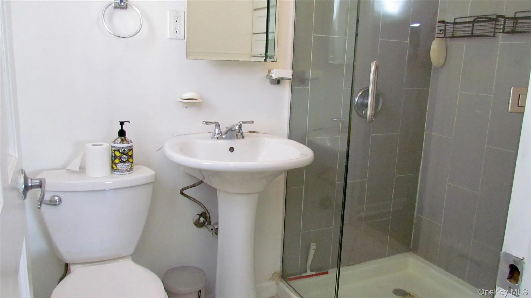 Bathroom featuring a pedestal sink with chrome faucet, toilet, and a glass-enclosed shower with gray tiled walls Bathroom featuring a pedestal sink with chrome faucet, toilet, and a glass-enclosed shower with gray tiled walls