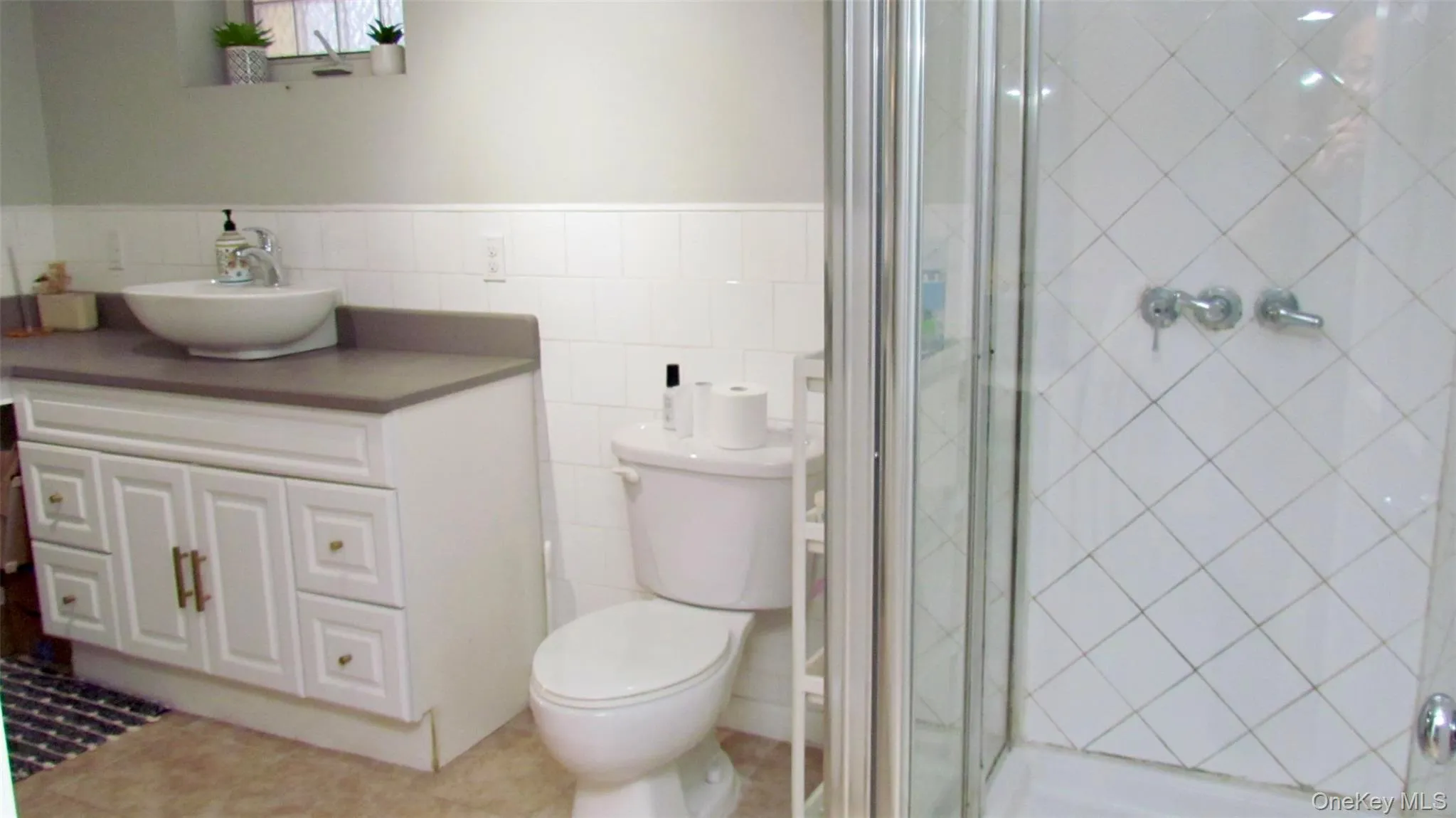 Bathroom featuring a walk-in shower with glass enclosure, white tile walls, a white vanity with vessel sink, and a toilet Bathroom featuring a walk-in shower with glass enclosure, white tile walls, a white vanity with vessel sink, and a toilet