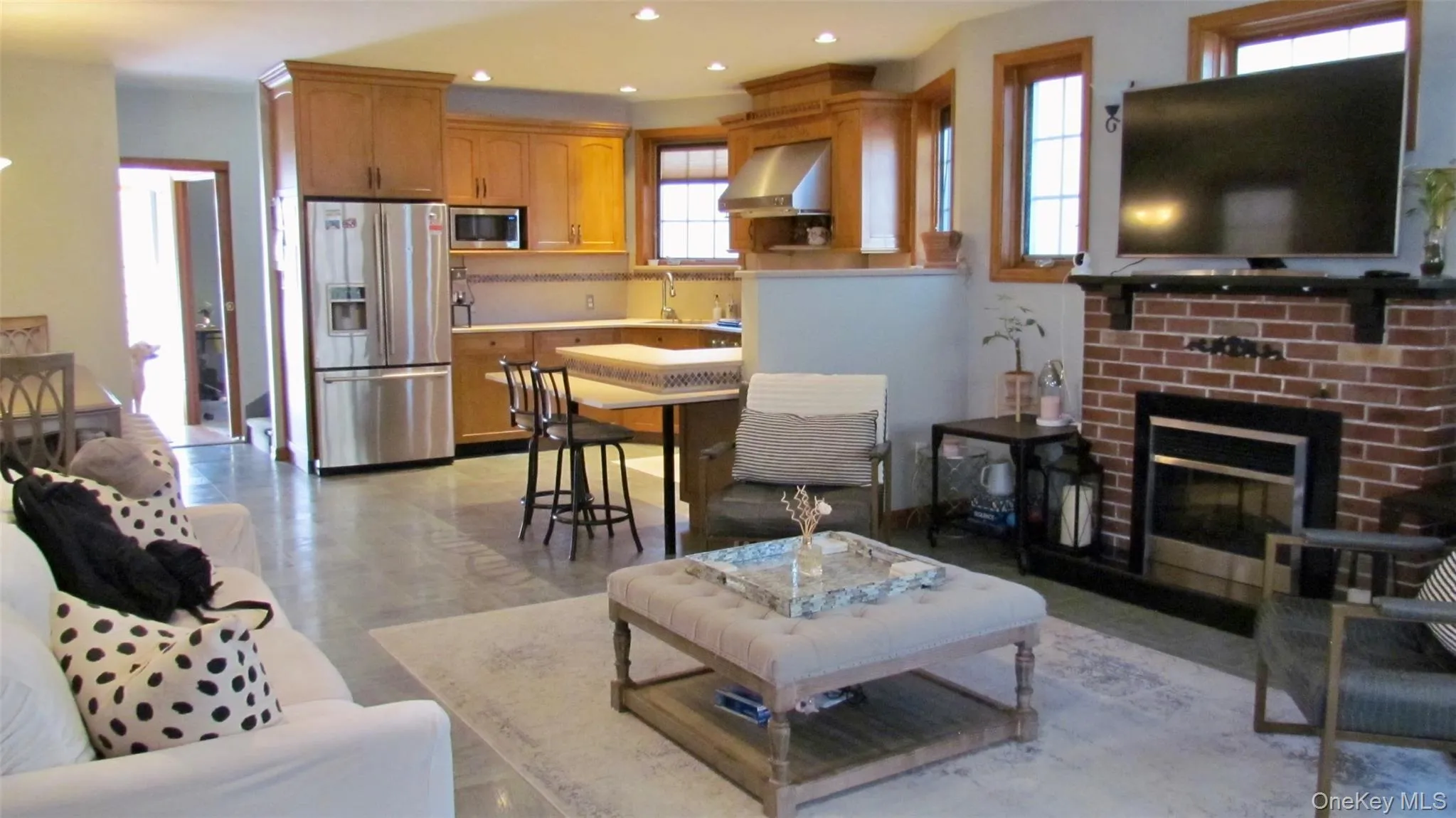 Open-concept living space featuring a brick fireplace with a dark mantel, wood-finish flooring, and natural wood window trim Open-concept living space featuring a brick fireplace with a dark mantel, wood-finish flooring, and natural wood window trim