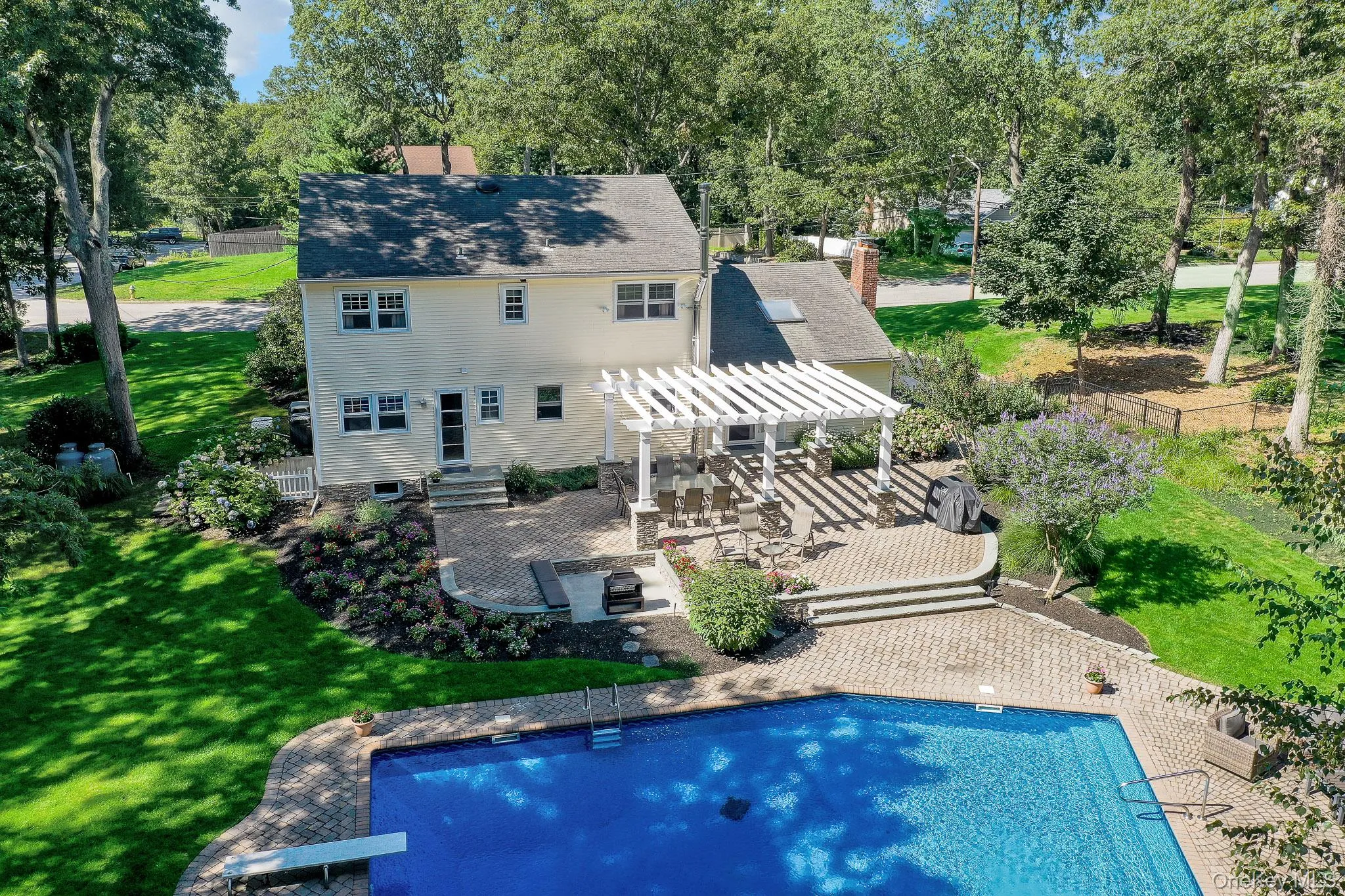 Expansive backyard featuring a rectangular swimming pool with a diving board, a large paver patio, and a white pergola covering an outdoor dining area Expansive backyard featuring a rectangular swimming pool with a diving board, a large paver patio, and a white pergola covering an outdoor dining area