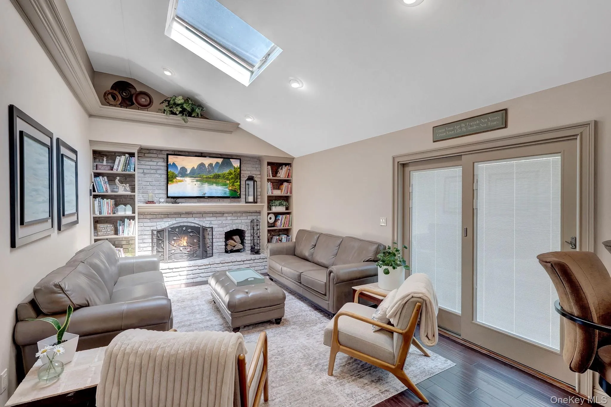 Vaulted ceiling living room featuring a white-painted brick fireplace, built-in shelving, a skylight, and dark wood-finish flooring Vaulted ceiling living room featuring a white-painted brick fireplace, built-in shelving, a skylight, and dark wood-finish flooring