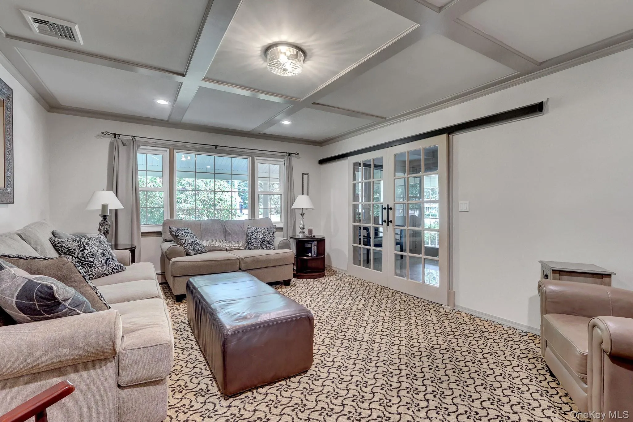 Living area featuring a coffered ceiling with recessed lighting and a central flush-mount fixture, patterned carpet flooring, a large window array with outdoor views, and glass-paned double doors on a barn door track, hidden hard wood floors Living area featuring a coffered ceiling with recessed lighting and a central flush-mount fixture, patterned carpet flooring, a large window array with outdoor views, and glass-paned double doors on a barn door track, hidden hard wood floors