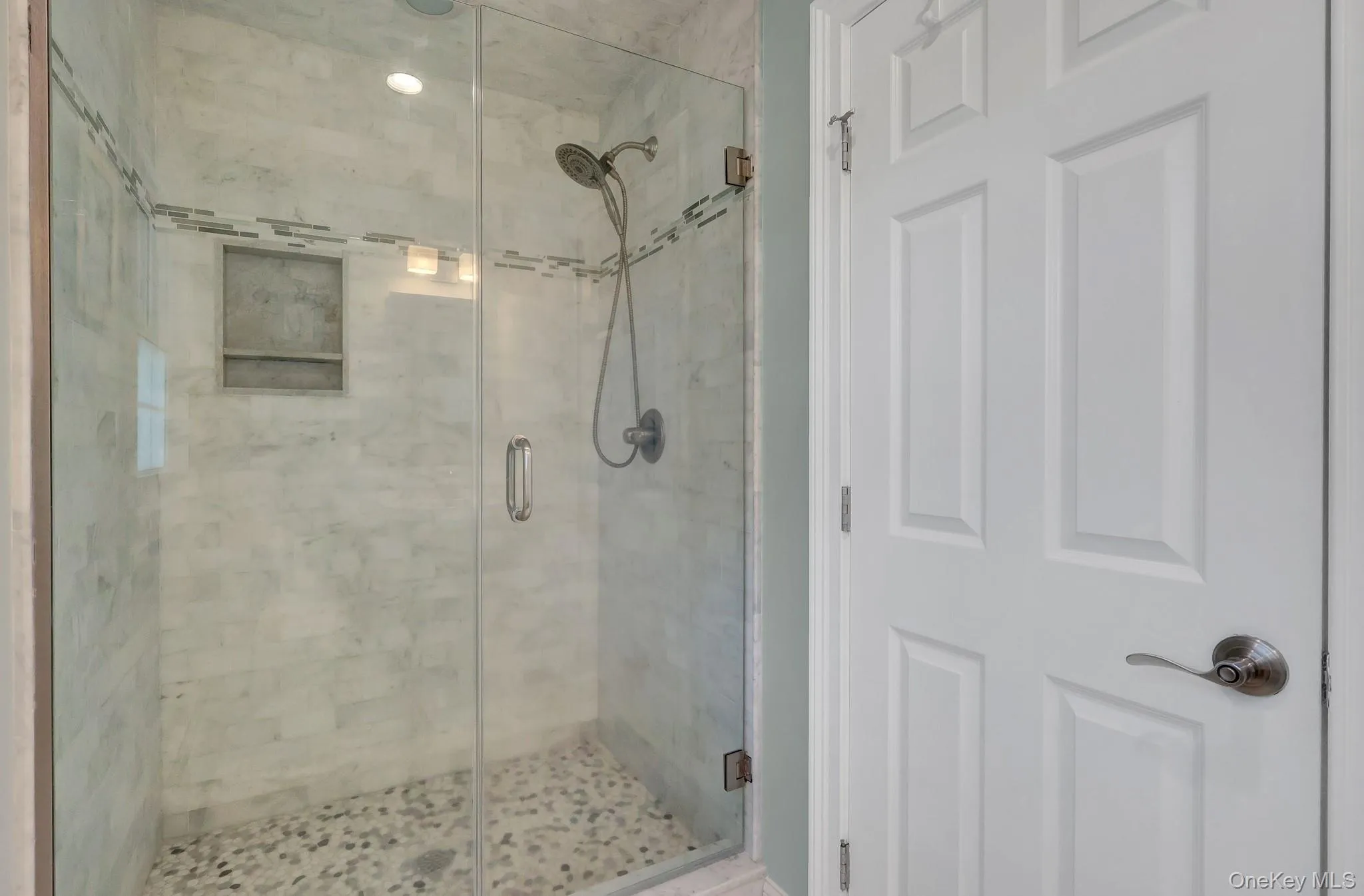 Glass-enclosed shower featuring marble-look wall tiling, decorative mosaic accent tile, a recessed shower niche, and pebble-finish flooring Glass-enclosed shower featuring marble-look wall tiling, decorative mosaic accent tile, a recessed shower niche, and pebble-finish flooring