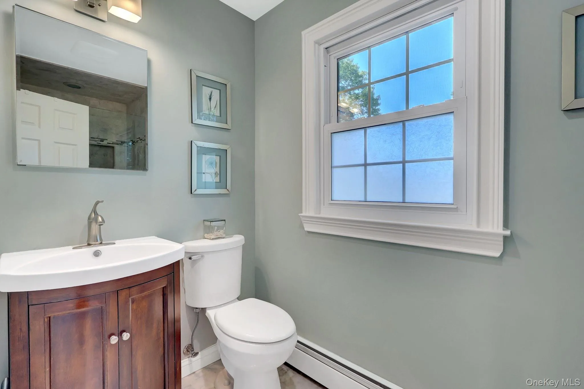 Bathroom featuring a wood-finish vanity with a white basin sink, brushed nickel faucet, and a wall-mounted mirror and radiant heat flooring. Bathroom featuring a wood-finish vanity with a white basin sink, brushed nickel faucet, and a wall-mounted mirror and radiant heat flooring.