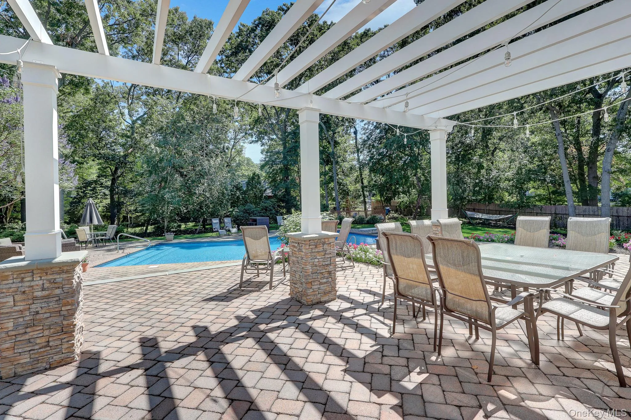 Expansive paver patio with a white pergola featuring string lighting and stone-clad pillars Expansive paver patio with a white pergola featuring string lighting and stone-clad pillars