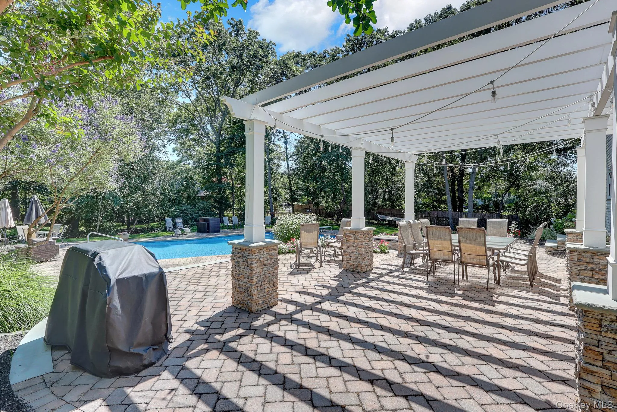 Expansive paver patio featuring a white pergola with string lighting, a swimming pool, and stone-clad pillars Expansive paver patio featuring a white pergola with string lighting, a swimming pool, and stone-clad pillars