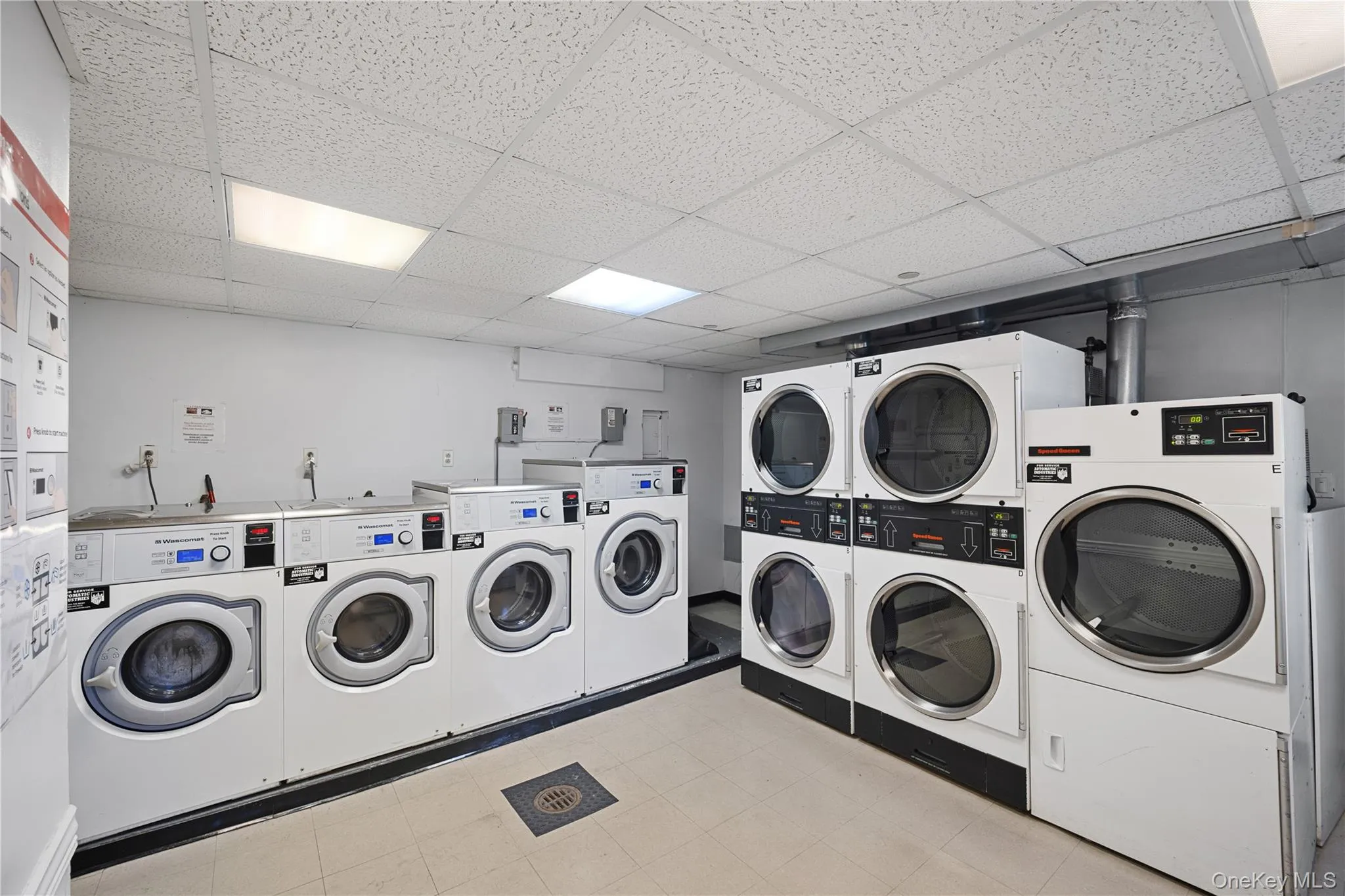 Dedicated laundry room featuring multiple commercial-grade front-loading washing machines and stacked dryers, overhead fluorescent lighting, a drop ceiling, and a floor drain Dedicated laundry room featuring multiple commercial-grade front-loading washing machines and stacked dryers, overhead fluorescent lighting, a drop ceiling, and a floor drain