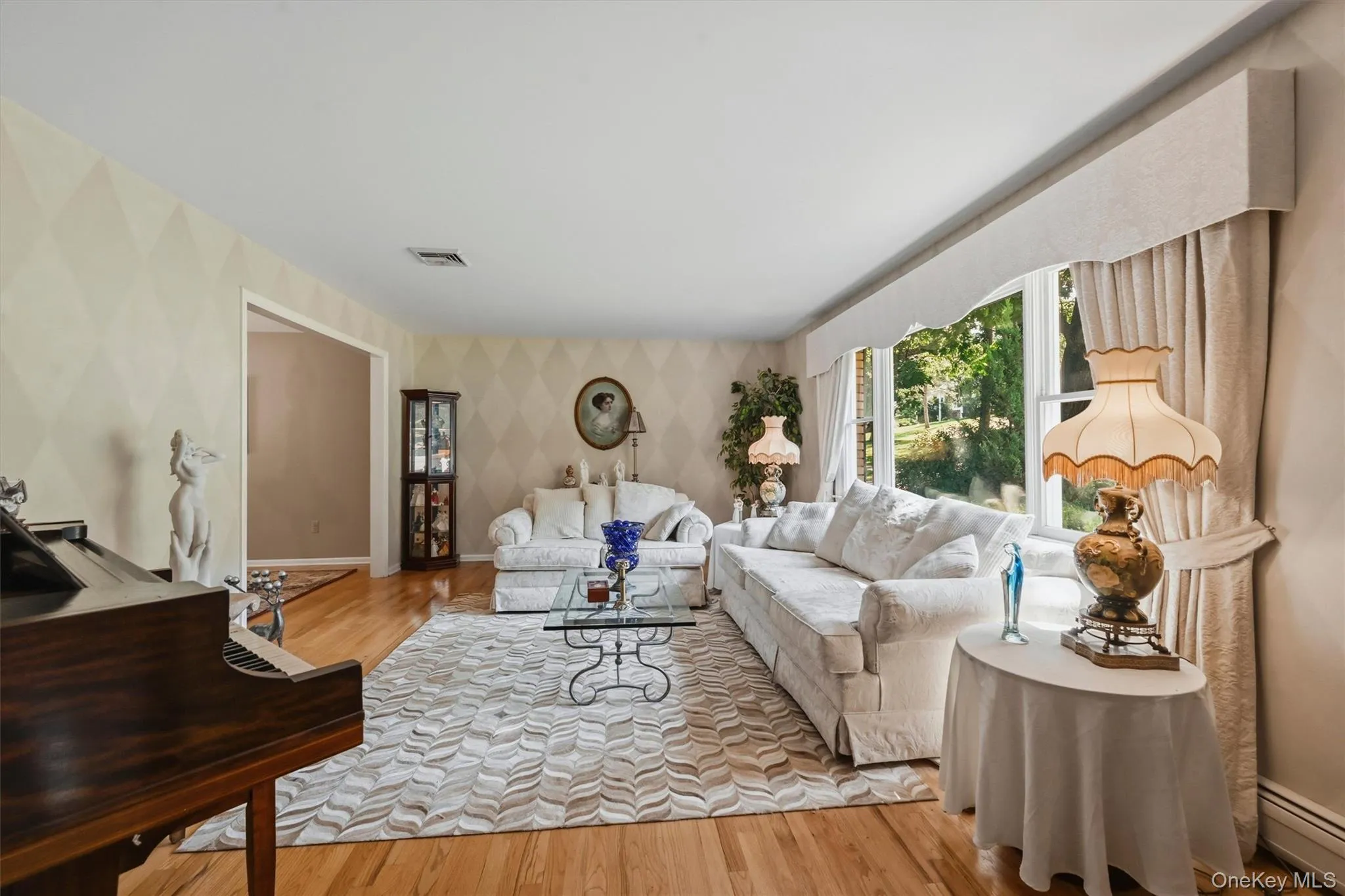 Living area featuring wood-finish flooring, patterned wallpaper, a large window with custom valance, and an open doorway Living area featuring wood-finish flooring, patterned wallpaper, a large window with custom valance, and an open doorway