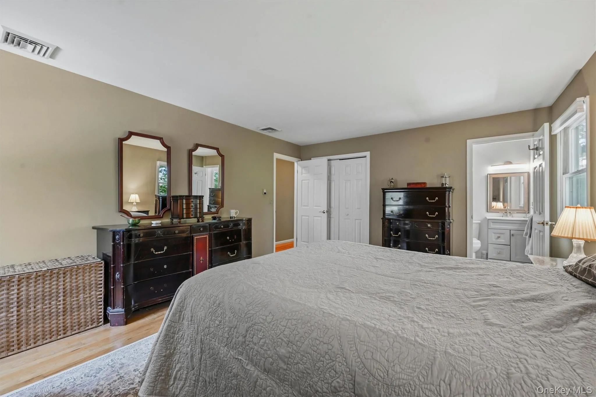 Spacious bedroom with light wood-finish flooring, neutral wall tones, white trim, and a private en-suite bathroom featuring a light gray vanity and mirrored medicine cabinet Spacious bedroom with light wood-finish flooring, neutral wall tones, white trim, and a private en-suite bathroom featuring a light gray vanity and mirrored medicine cabinet