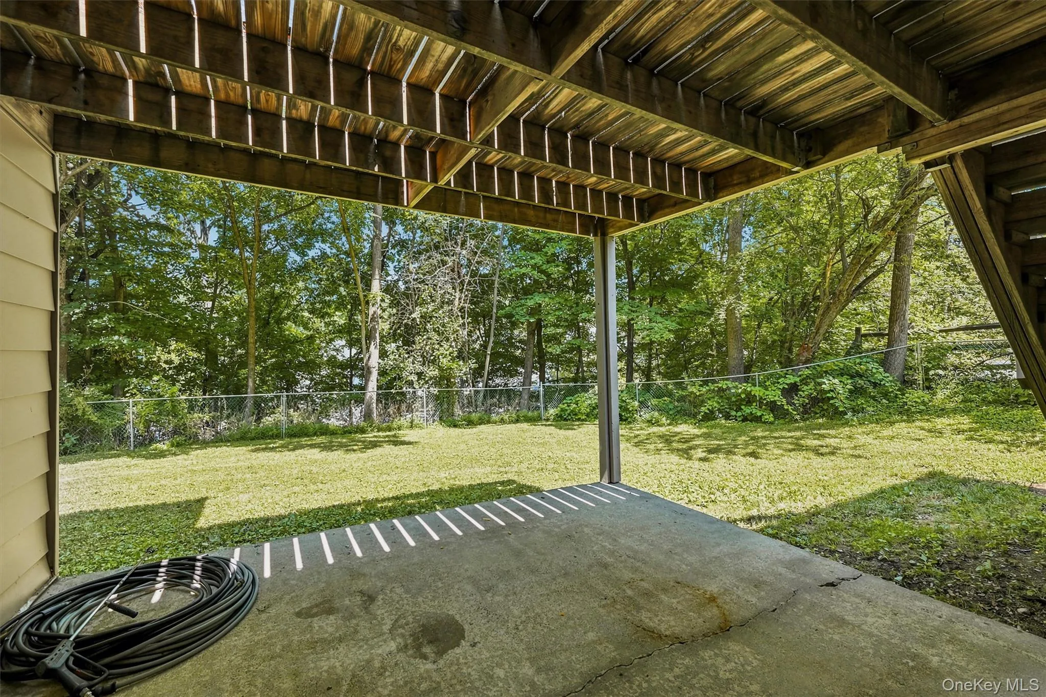 Covered concrete patio with wooden overhead structure and support posts, opening onto a fenced lawn area with mature trees Covered concrete patio with wooden overhead structure and support posts, opening onto a fenced lawn area with mature trees
