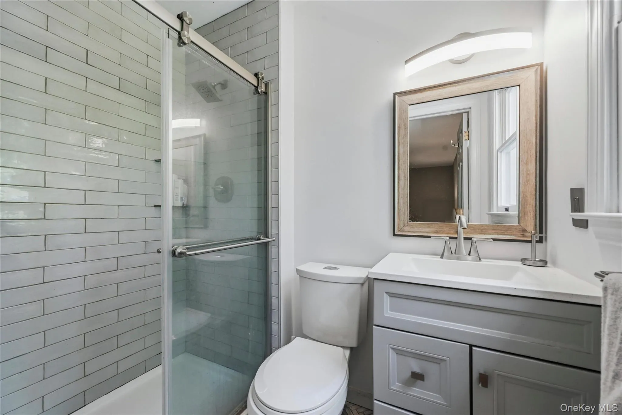 Bathroom featuring a glass-enclosed shower with subway tile surround, gray vanity with integrated sink, framed mirror, and contemporary light fixture Bathroom featuring a glass-enclosed shower with subway tile surround, gray vanity with integrated sink, framed mirror, and contemporary light fixture