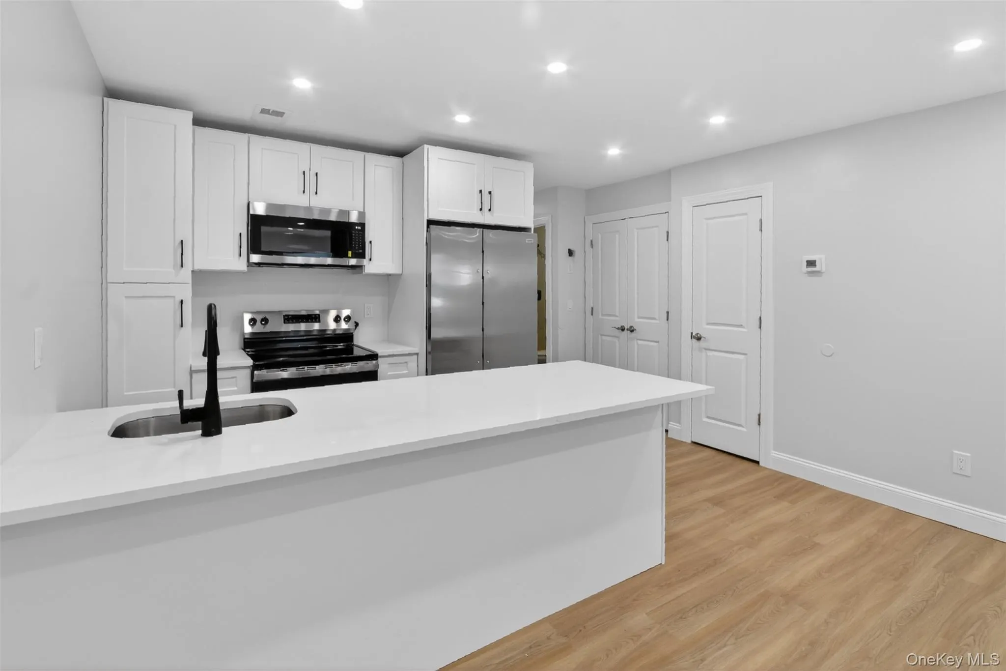 Contemporary kitchen featuring a large island with an undermount sink and matte black faucet, white shaker cabinetry, stainless steel appliances, and recessed lighting Contemporary kitchen featuring a large island with an undermount sink and matte black faucet, white shaker cabinetry, stainless steel appliances, and recessed lighting