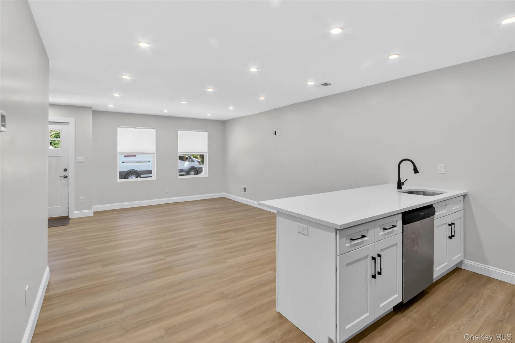 Open-concept area featuring light wood-finish flooring, recessed lighting, and a kitchen island with a built-in sink, stainless steel dishwasher, and white cabinetry with dark hardware Open-concept area featuring light wood-finish flooring, recessed lighting, and a kitchen island with a built-in sink, stainless steel dishwasher, and white cabinetry with dark hardware