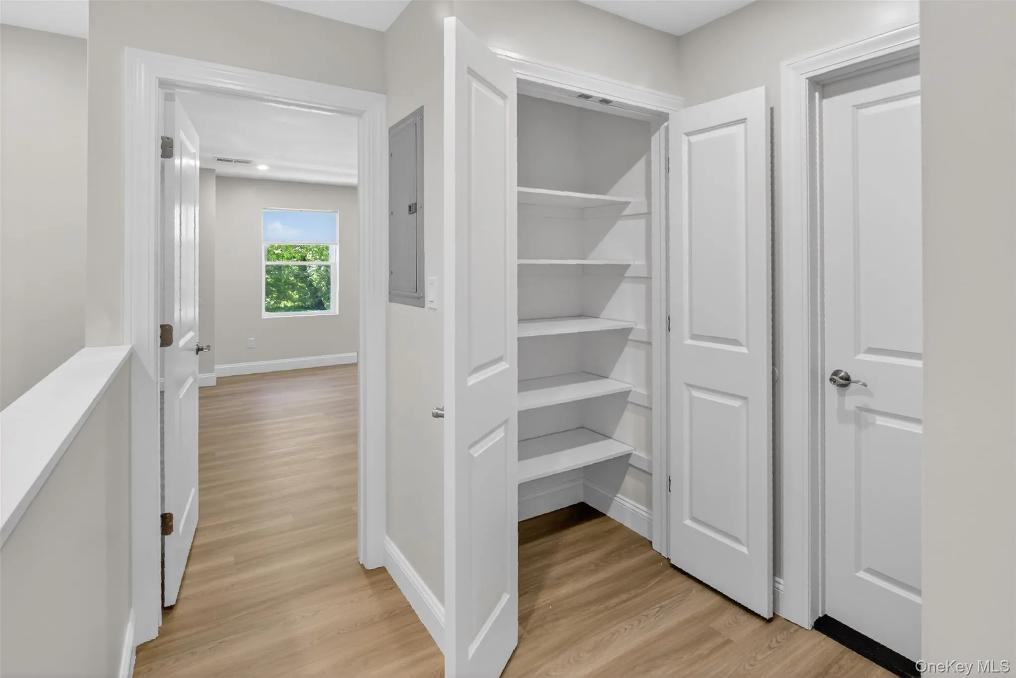 Hallway with wood-finish flooring, featuring a built-in pantry with shelving, white paneled doors, and light gray wall paint Hallway with wood-finish flooring, featuring a built-in pantry with shelving, white paneled doors, and light gray wall paint