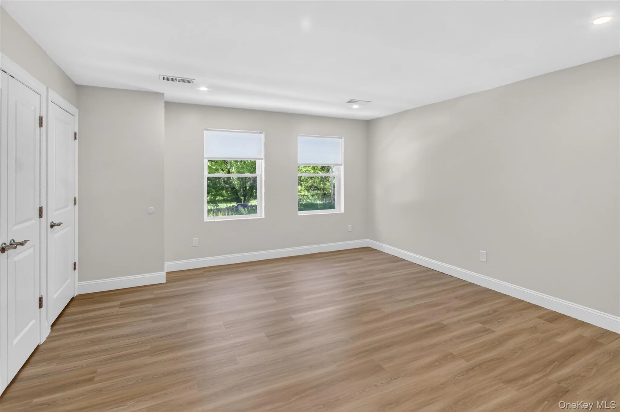 Spacious room featuring light-toned wood-finish flooring, two large windows, white baseboards, recessed lighting, and a neutral wall color Spacious room featuring light-toned wood-finish flooring, two large windows, white baseboards, recessed lighting, and a neutral wall color