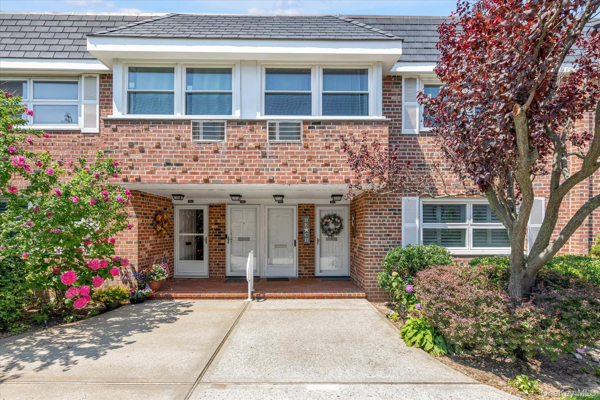 Brick facade townhome featuring white window shutters, a dark shingle roof, and mature landscaping Brick facade townhome featuring white window shutters, a dark shingle roof, and mature landscaping