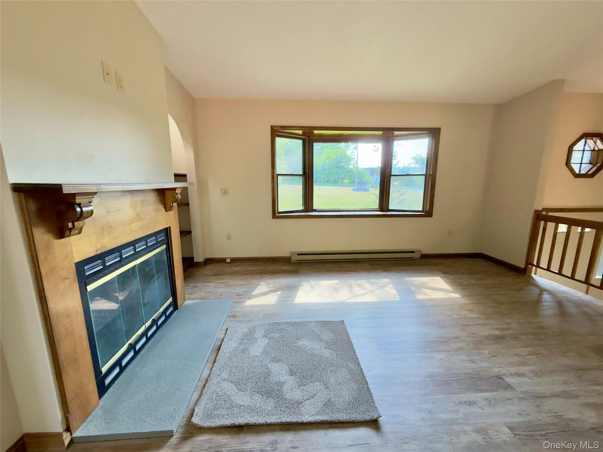 Living area featuring a wood-surround fireplace with a gray hearth, a bay window, wood-finish flooring, and a natural wood railing Living area featuring a wood-surround fireplace with a gray hearth, a bay window, wood-finish flooring, and a natural wood railing