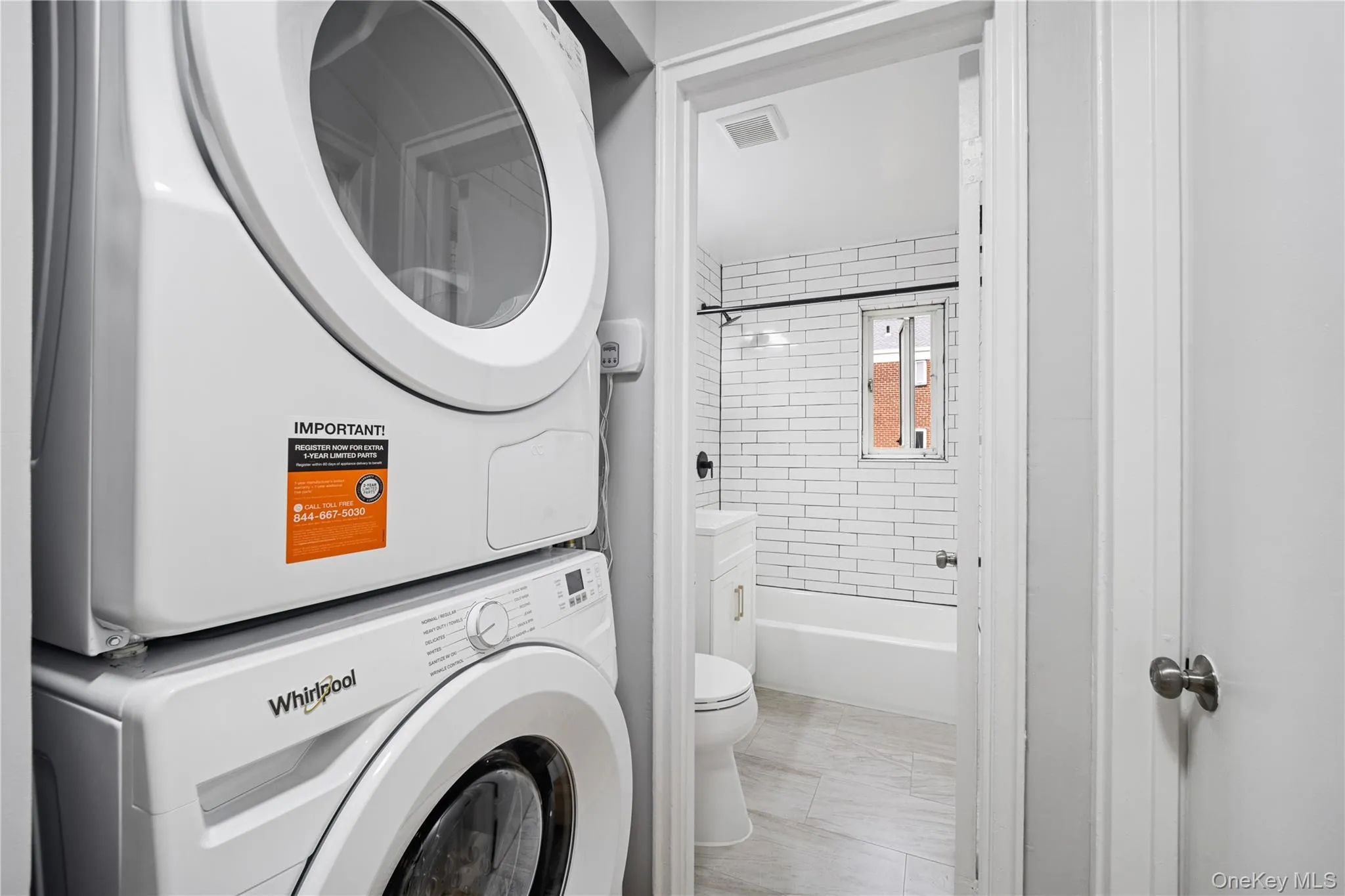 Compact laundry area featuring a stacked washer and dryer, adjacent to a bathroom with subway tile shower surround, a bathtub, and a window Compact laundry area featuring a stacked washer and dryer, adjacent to a bathroom with subway tile shower surround, a bathtub, and a window