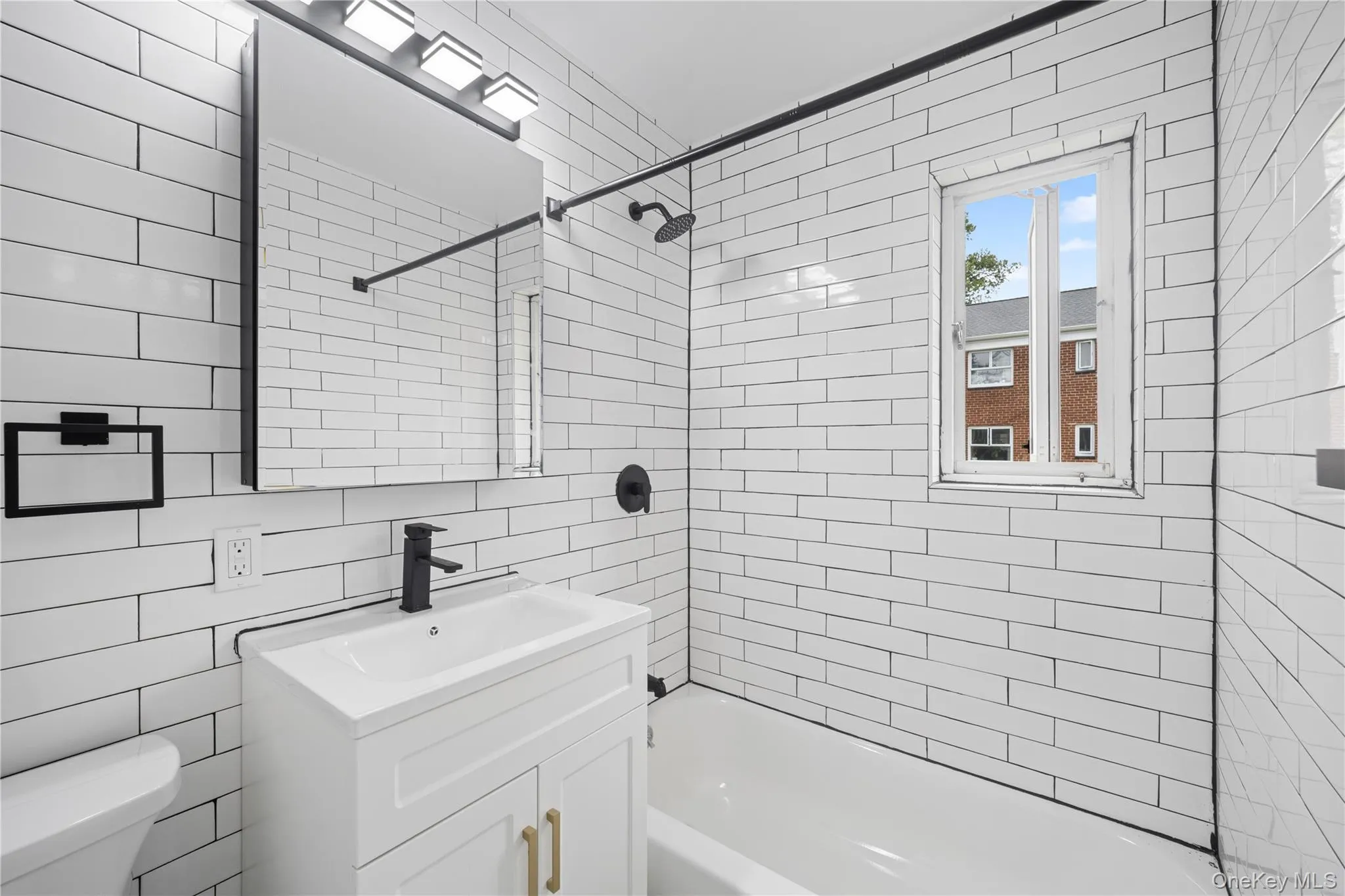 Bathroom featuring white subway tile surround, matte black fixtures, a white vanity with gold-tone hardware, and a window Bathroom featuring white subway tile surround, matte black fixtures, a white vanity with gold-tone hardware, and a window