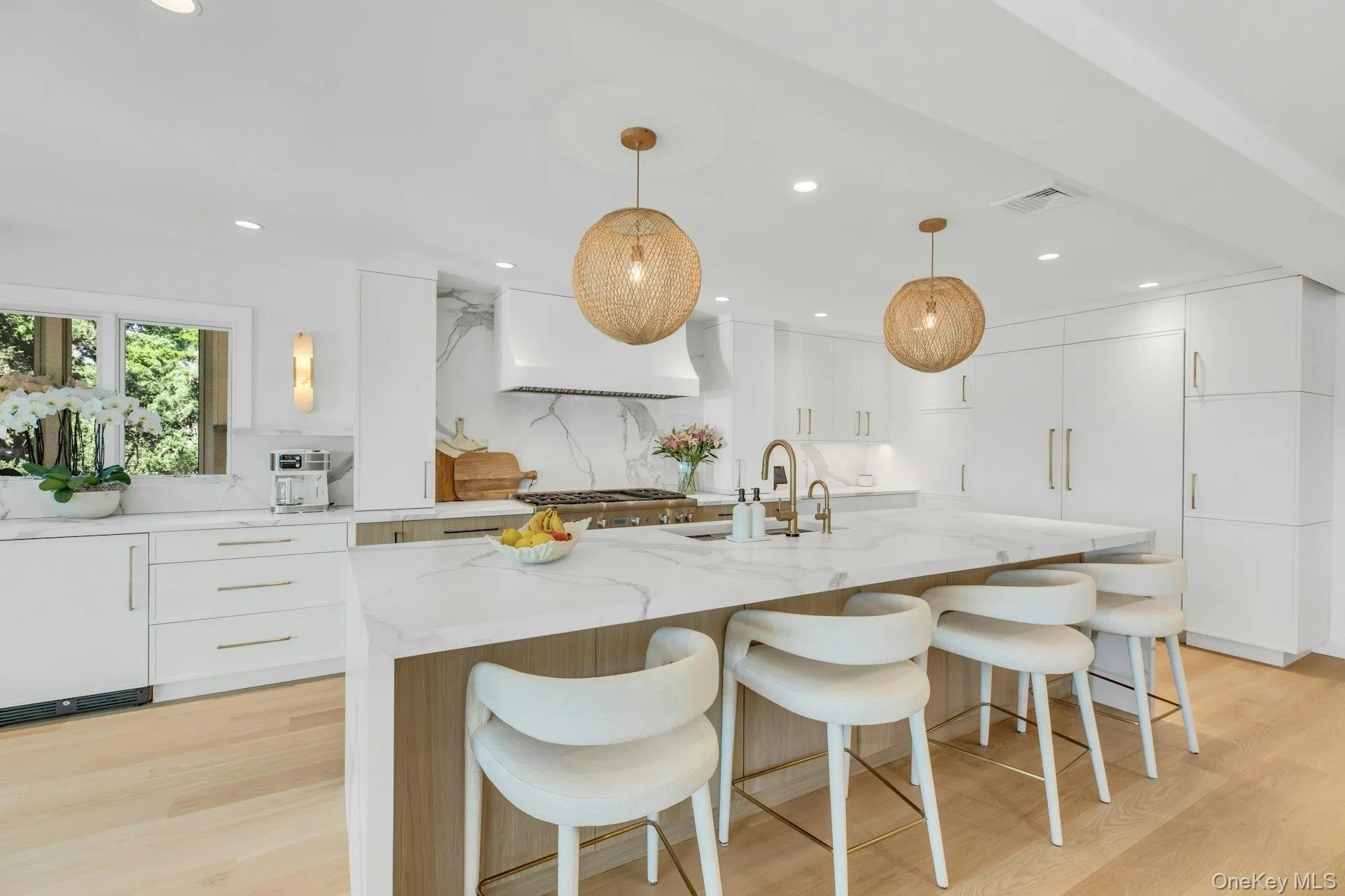 Spacious kitchen featuring an oversized island with countertop seating, light wood-finish flooring, white cabinetry with gold-tone hardware, and integrated appliances Spacious kitchen featuring an oversized island with countertop seating, light wood-finish flooring, white cabinetry with gold-tone hardware, and integrated appliances