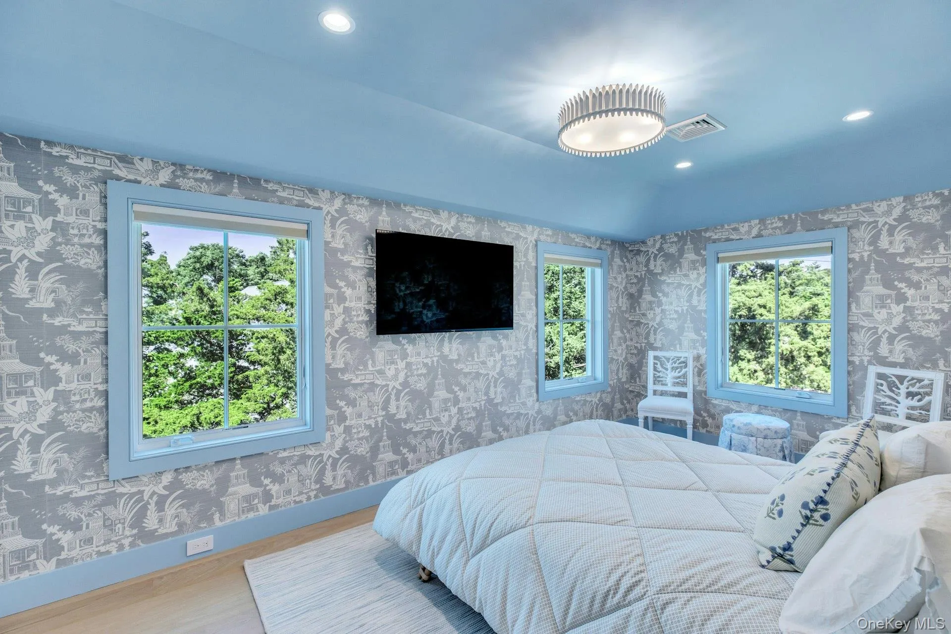 Spacious bedroom featuring light wood-finish flooring, patterned wallpaper, a built-in wall-mounted television, and multiple windows with light blue trim Spacious bedroom featuring light wood-finish flooring, patterned wallpaper, a built-in wall-mounted television, and multiple windows with light blue trim