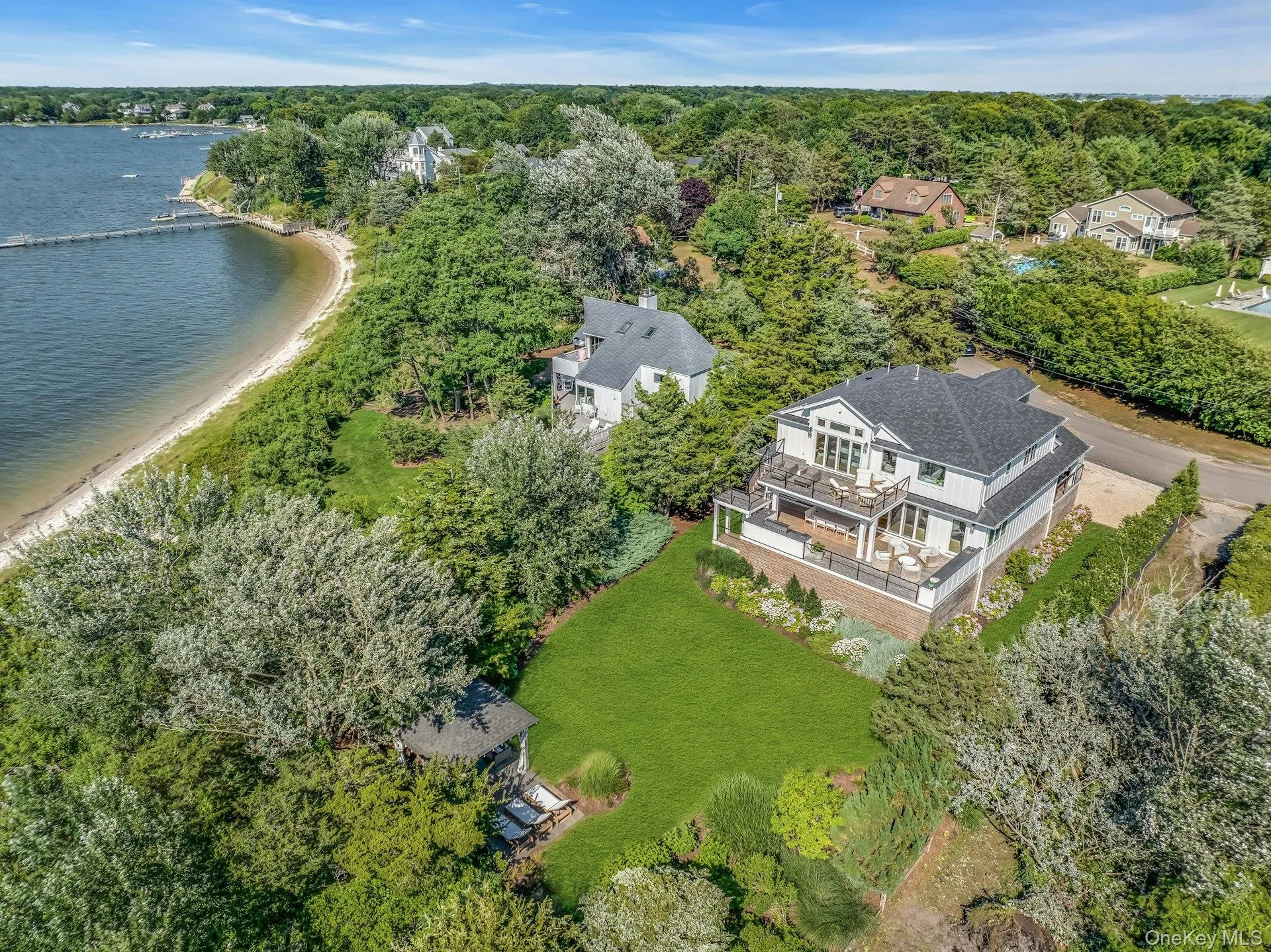 Modern coastal home with multiple outdoor decks, a manicured lawn, and mature trees, featuring a private sandy beach and a long dock extending into the water Modern coastal home with multiple outdoor decks, a manicured lawn, and mature trees, featuring a private sandy beach and a long dock extending into the water
