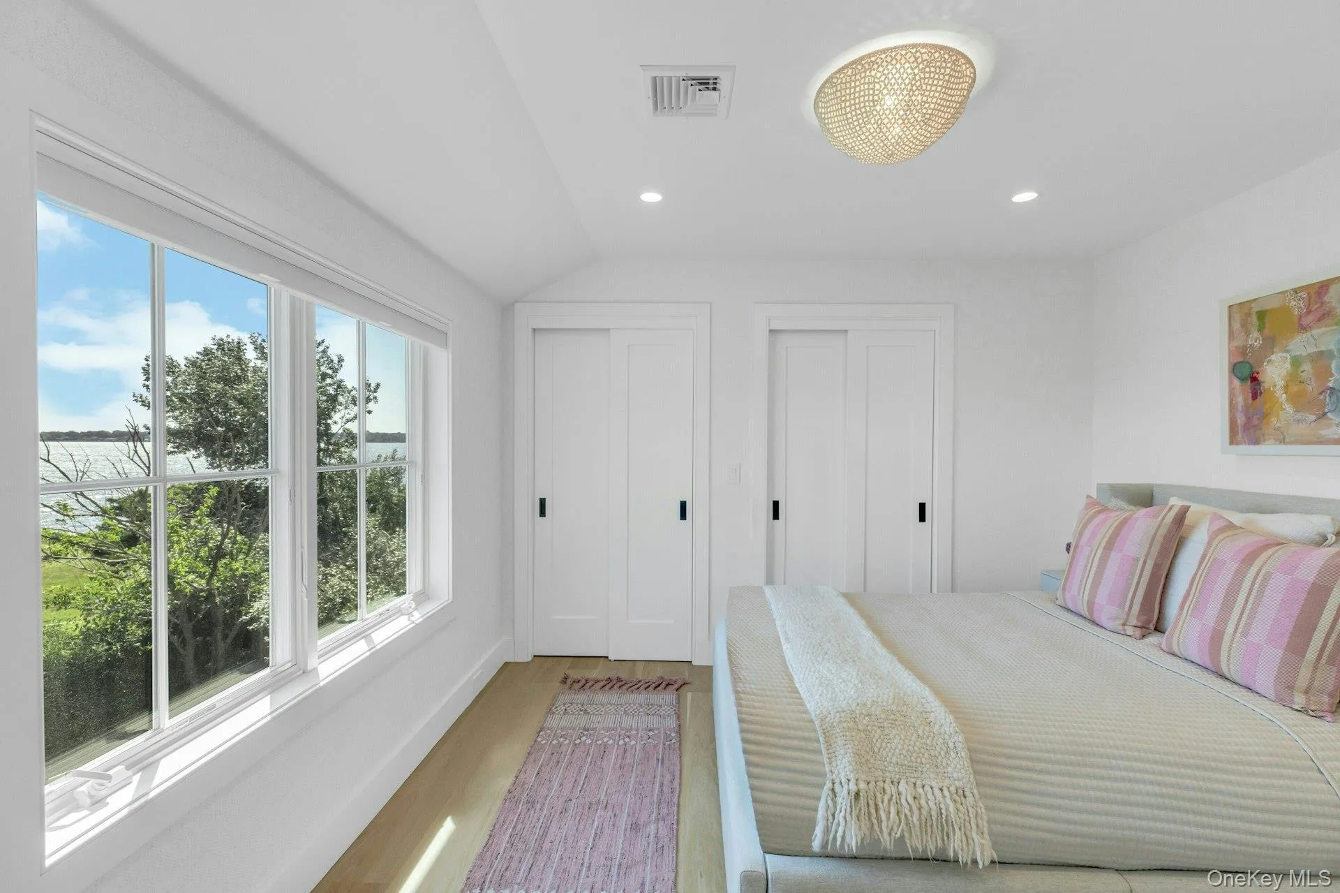 Light-filled bedroom featuring a multi-pane window overlooking a body of water and lush greenery, wood-finish flooring, two sliding closet doors with dark hardware, a flush-mount ceiling light fixture, and recessed lighting Light-filled bedroom featuring a multi-pane window overlooking a body of water and lush greenery, wood-finish flooring, two sliding closet doors with dark hardware, a flush-mount ceiling light fixture, and recessed lighting