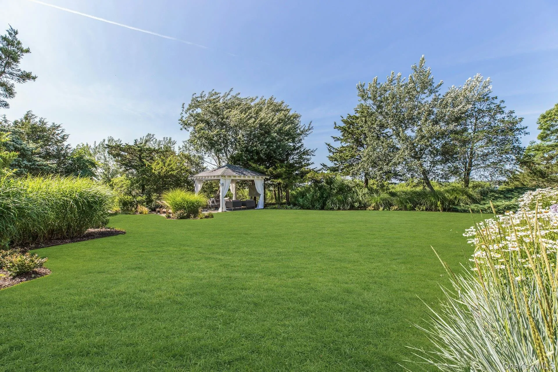 Expansive manicured lawn featuring a white gazebo with drapes, surrounded by mature trees and varied landscaping Expansive manicured lawn featuring a white gazebo with drapes, surrounded by mature trees and varied landscaping