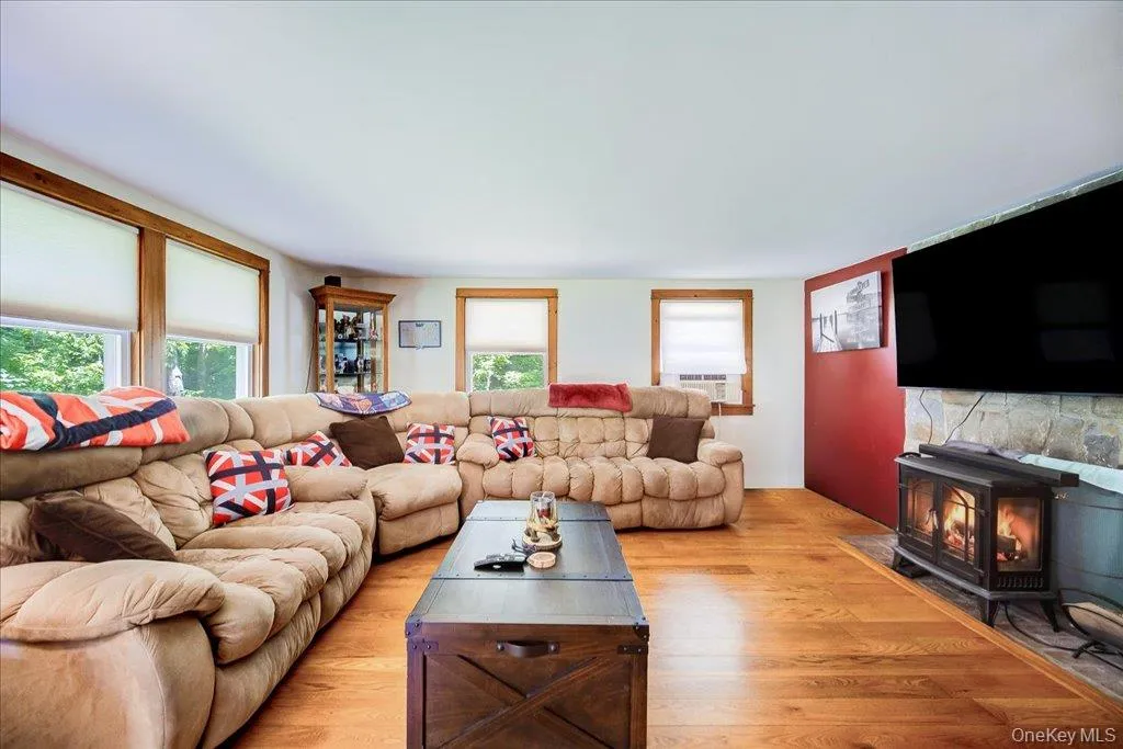 Living area featuring wood-finish flooring, multiple windows with natural wood trim, a stone-faced hearth, and a freestanding fireplace Living area featuring wood-finish flooring, multiple windows with natural wood trim, a stone-faced hearth, and a freestanding fireplace