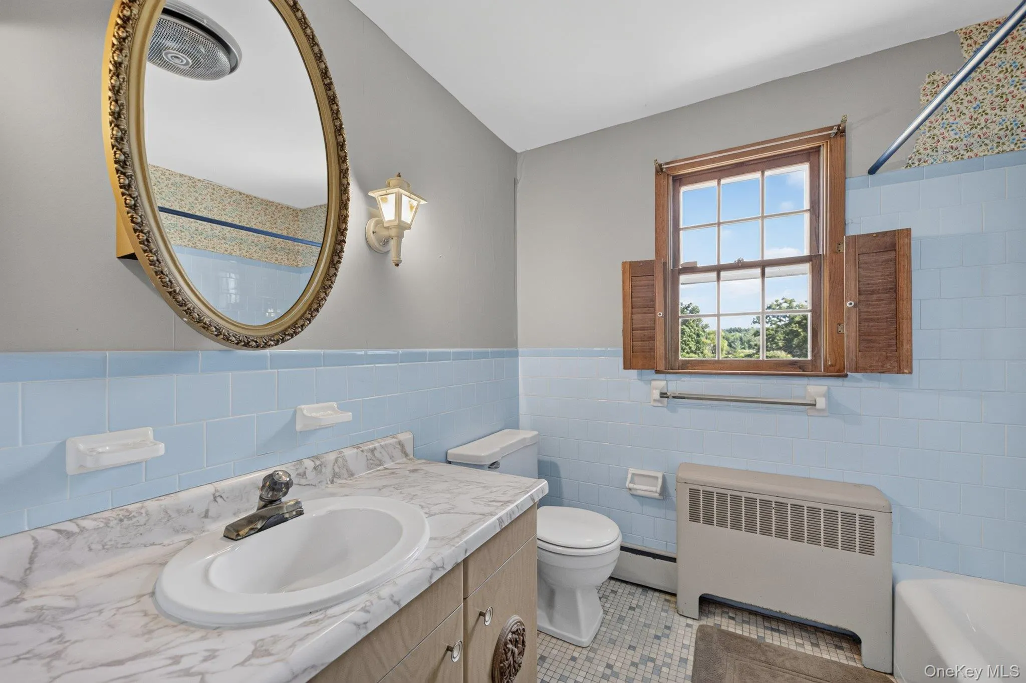 Bathroom featuring a vanity with a white countertop and integrated sink, an oval mirror with an ornate frame, a window with wood shutters, and blue wall tiles Bathroom featuring a vanity with a white countertop and integrated sink, an oval mirror with an ornate frame, a window with wood shutters, and blue wall tiles