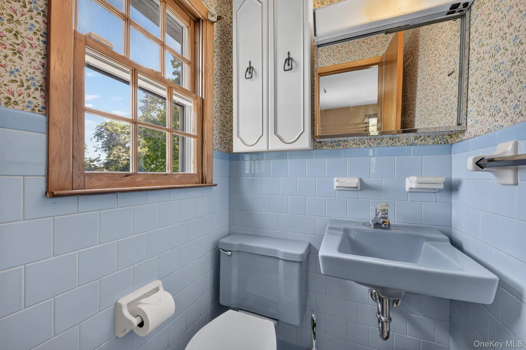 Bathroom featuring light blue wall tiles, a wood-framed window, a wall-mounted sink, and a mirrored medicine cabinet Bathroom featuring light blue wall tiles, a wood-framed window, a wall-mounted sink, and a mirrored medicine cabinet