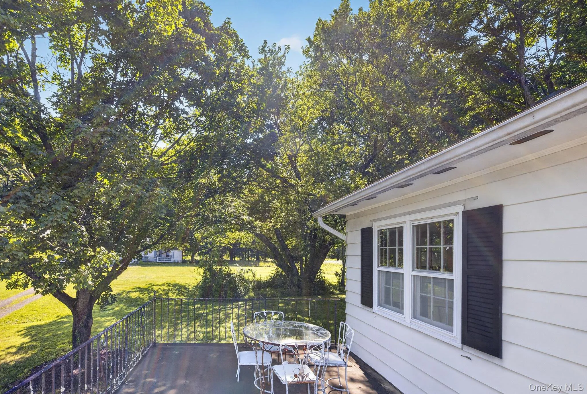White siding exterior with a dark-colored balcony railing, offering views of mature trees and a grassy expanse White siding exterior with a dark-colored balcony railing, offering views of mature trees and a grassy expanse