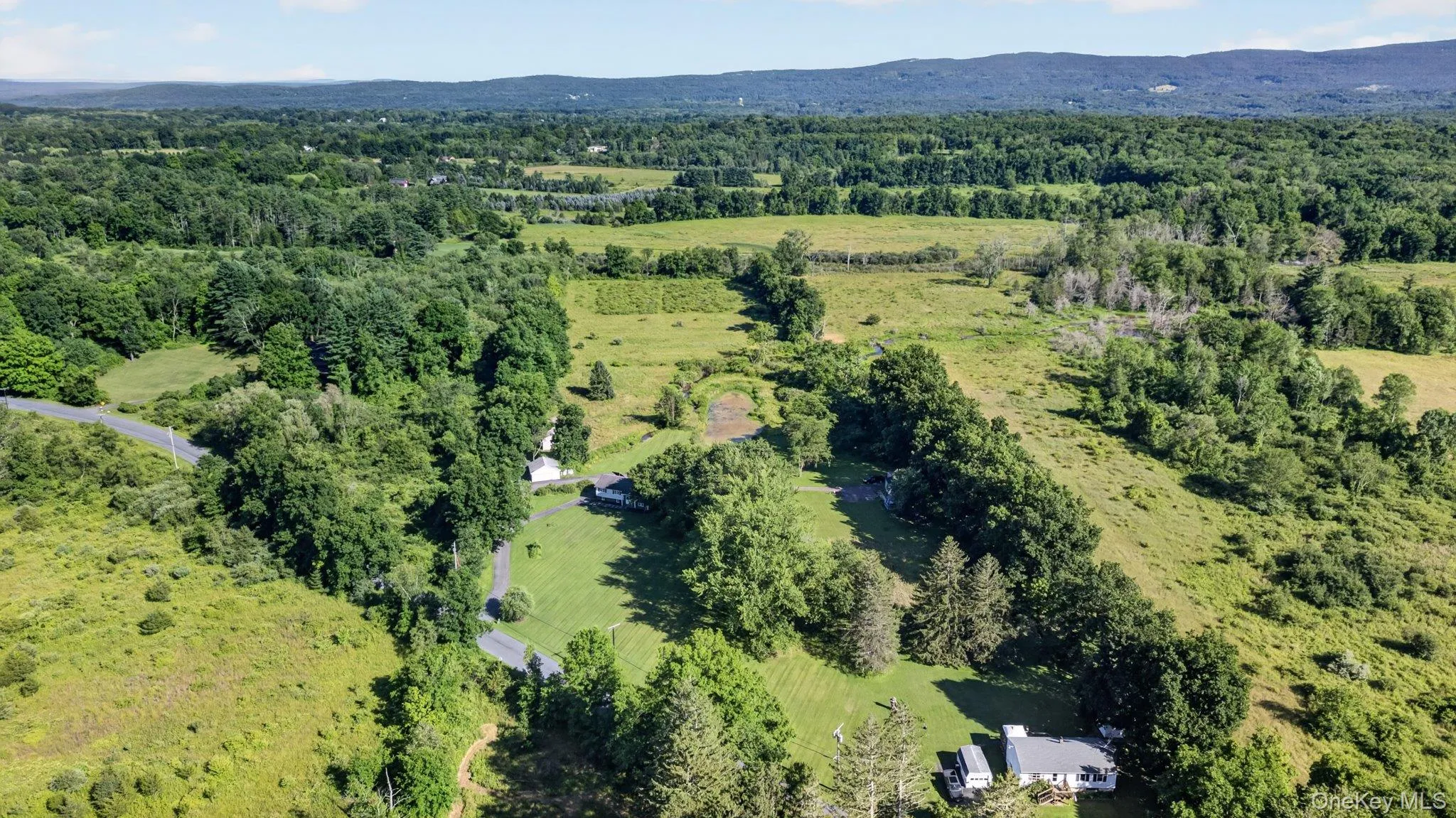 Expansive rural landscape featuring a winding paved road, multiple structures, mature trees, and distant mountainous terrain Expansive rural landscape featuring a winding paved road, multiple structures, mature trees, and distant mountainous terrain