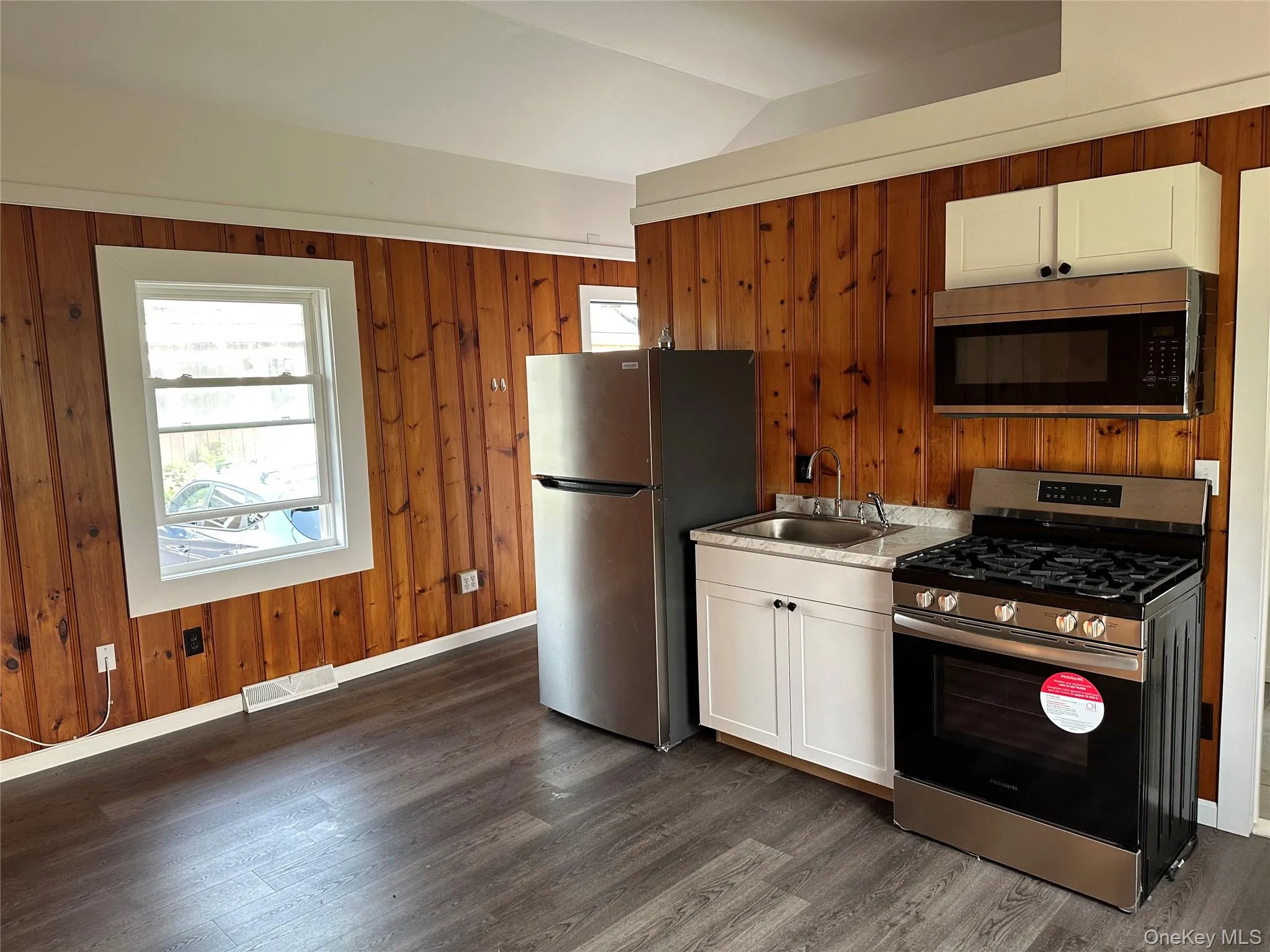 Compact kitchen area featuring wood-panel walls, stainless steel appliances, white cabinetry, and wood-finish flooring Compact kitchen area featuring wood-panel walls, stainless steel appliances, white cabinetry, and wood-finish flooring