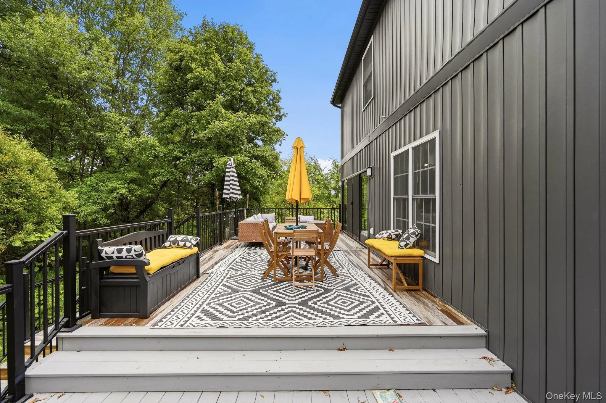 Expansive outdoor deck featuring wood-finish flooring, dark board-and-batten siding, black railings, and a mature tree canopy backdrop Expansive outdoor deck featuring wood-finish flooring, dark board-and-batten siding, black railings, and a mature tree canopy backdrop