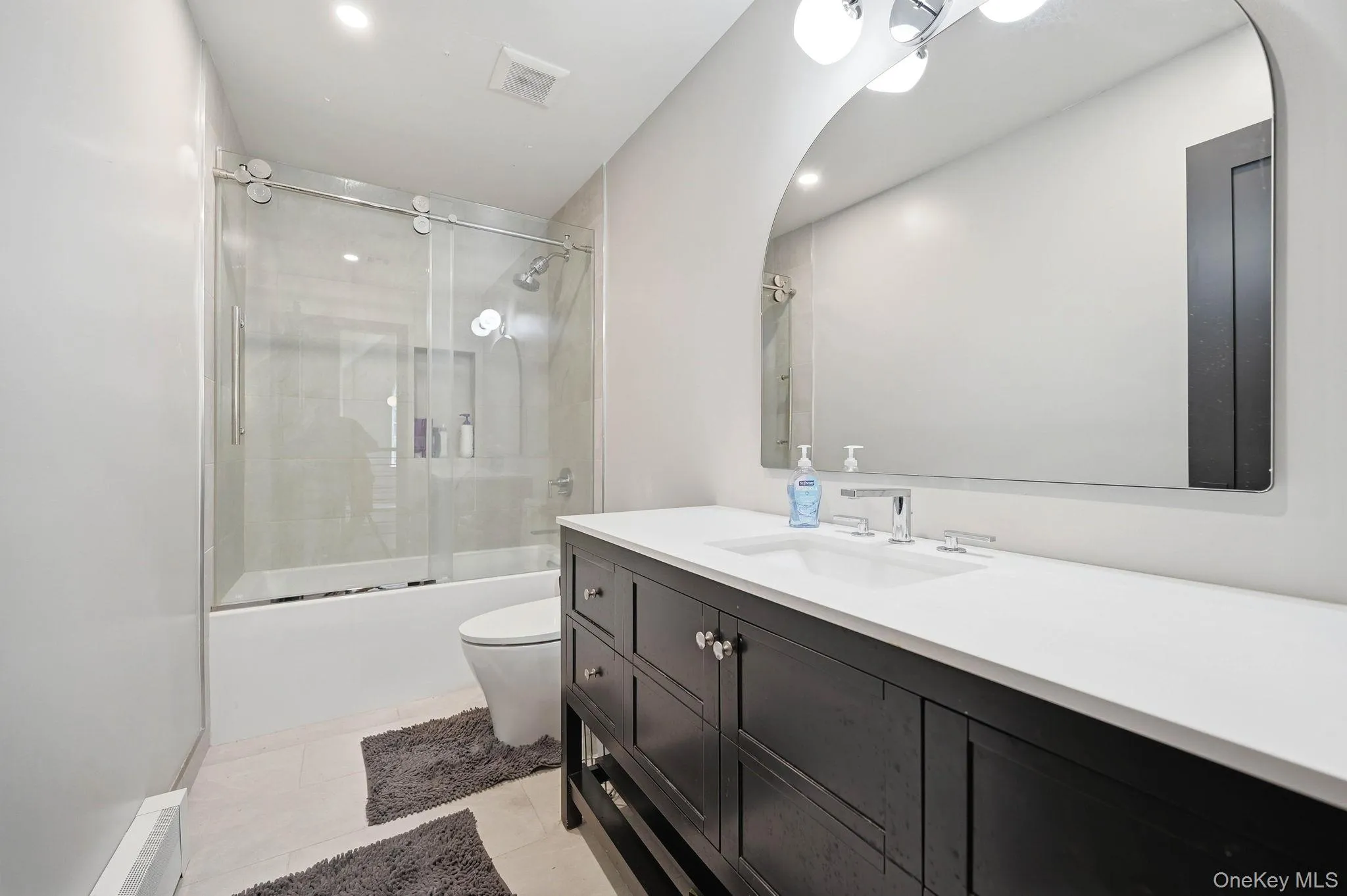 Modern bathroom featuring a dark wood-finish vanity with a white countertop and undermount sink, a large arched mirror, and a tub/shower combination with a sliding glass enclosure Modern bathroom featuring a dark wood-finish vanity with a white countertop and undermount sink, a large arched mirror, and a tub/shower combination with a sliding glass enclosure
