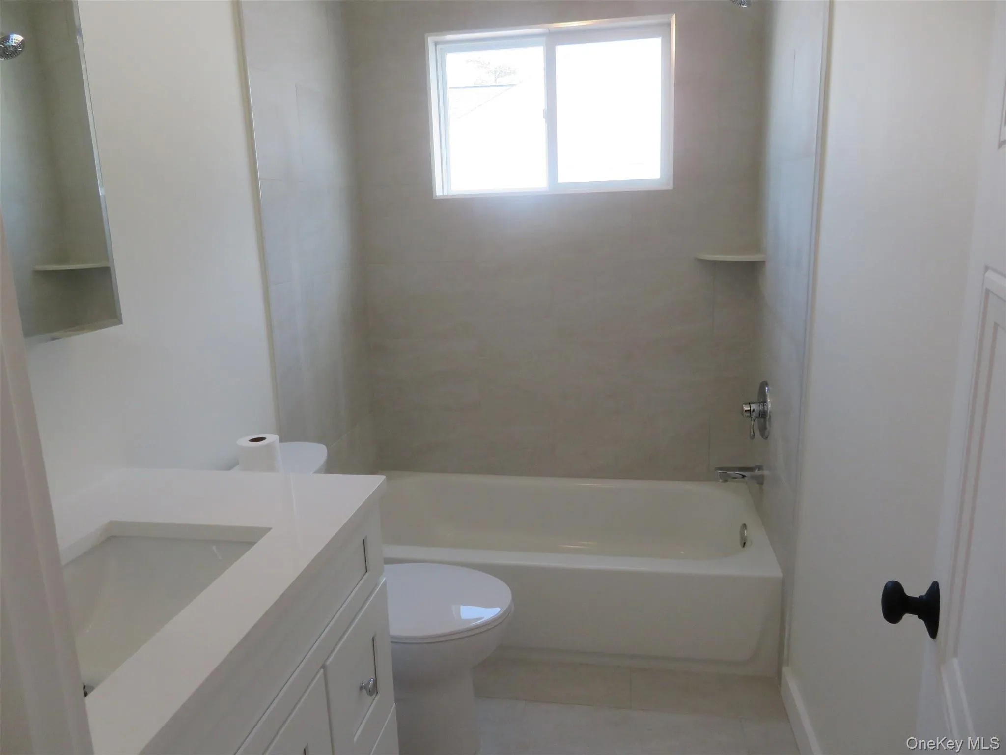 Bathroom featuring a white vanity with an integrated sink, a built-in bathtub with a shower, a tiled shower surround, a window, and a wall-mounted medicine cabinet Bathroom featuring a white vanity with an integrated sink, a built-in bathtub with a shower, a tiled shower surround, a window, and a wall-mounted medicine cabinet