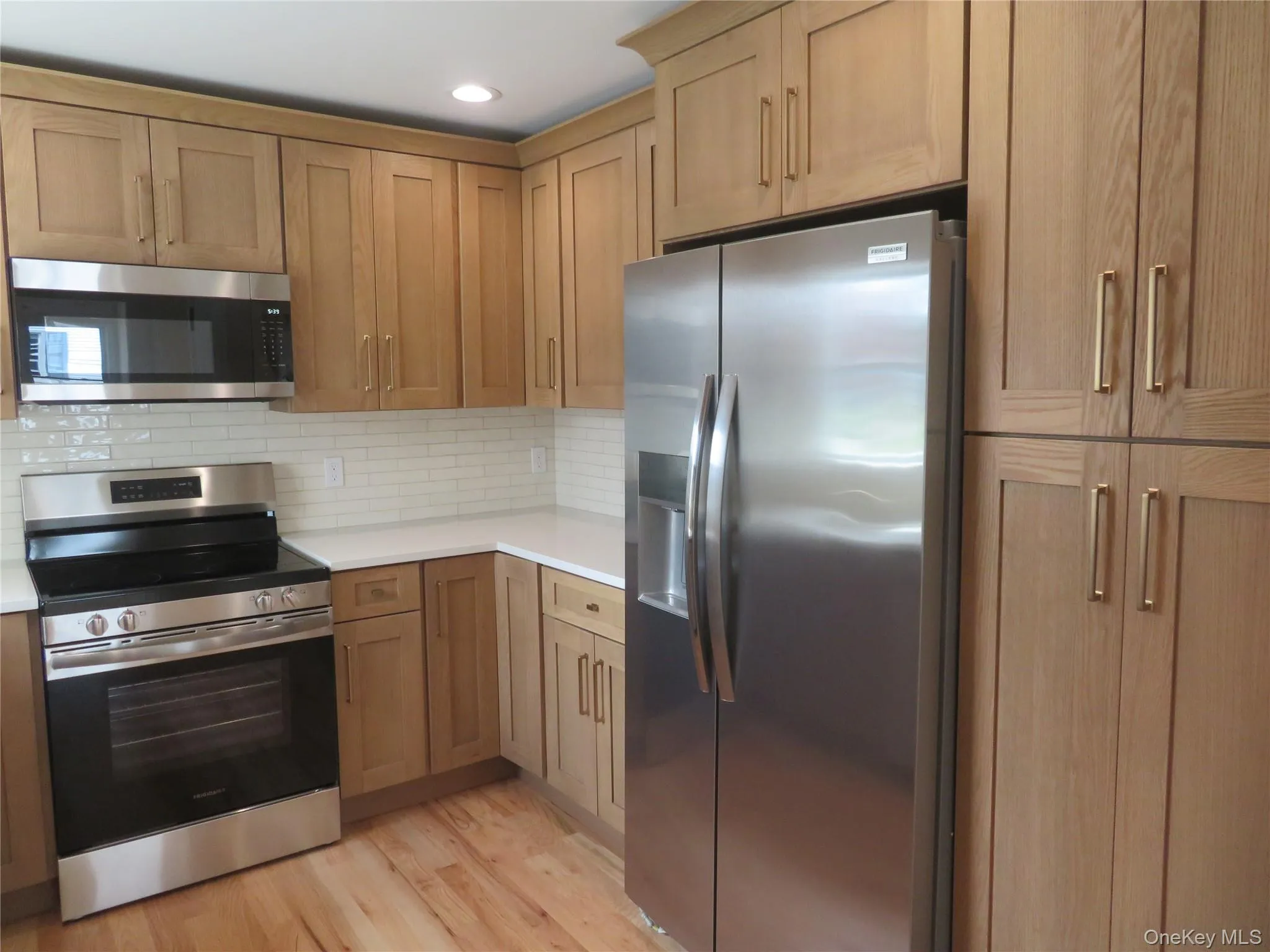 Kitchen featuring light wood-finish cabinetry, stainless steel appliances, white subway tile backsplash, light-toned countertops, and wood-finish flooring Kitchen featuring light wood-finish cabinetry, stainless steel appliances, white subway tile backsplash, light-toned countertops, and wood-finish flooring