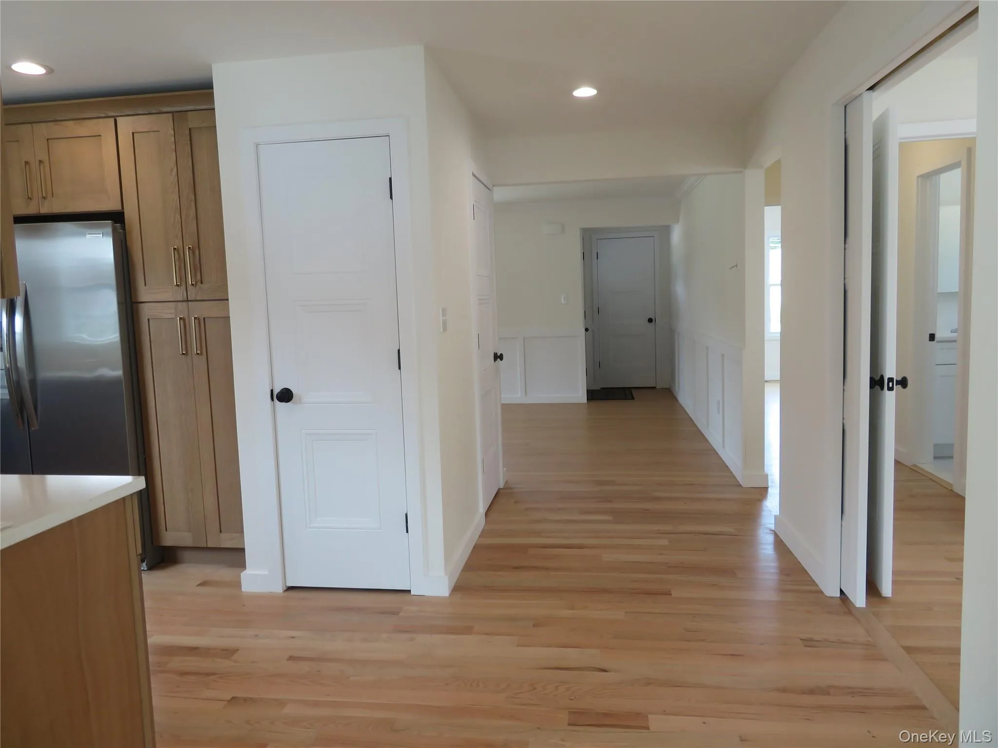 Open-concept space featuring wood-finish flooring, white walls, recessed lighting, and a kitchen area with light wood cabinetry and a stainless steel refrigerator Open-concept space featuring wood-finish flooring, white walls, recessed lighting, and a kitchen area with light wood cabinetry and a stainless steel refrigerator