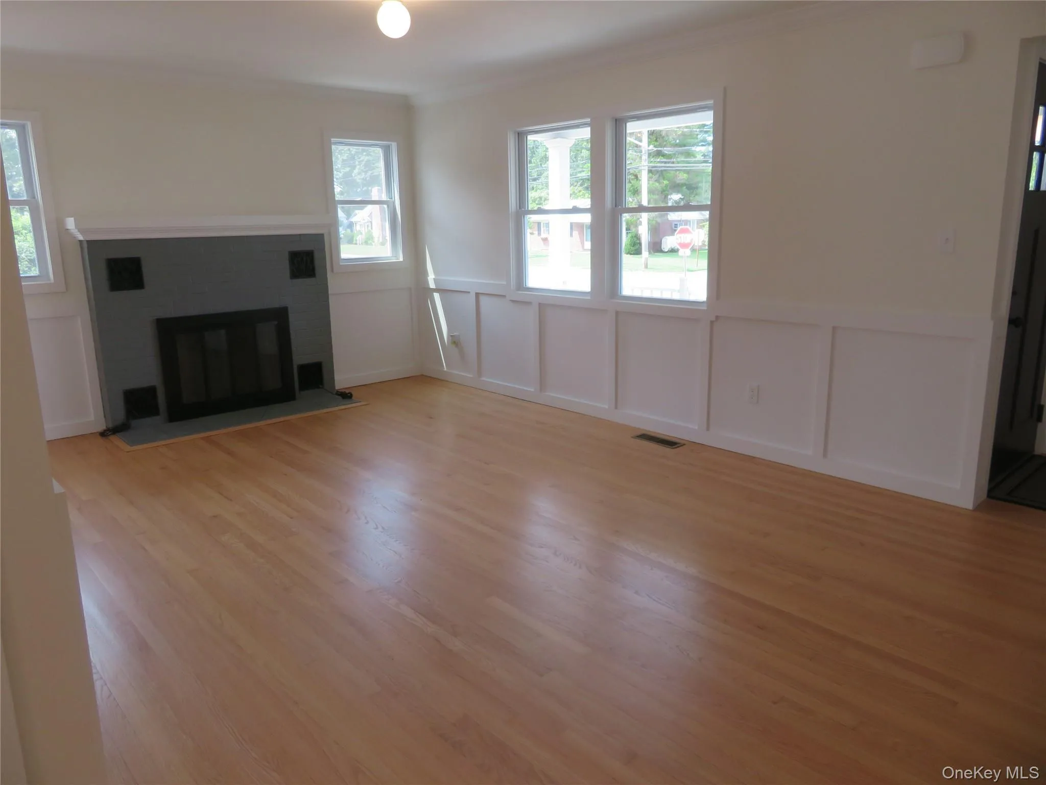 Living space featuring wood-finish flooring, a fireplace with a gray surround, white wainscoting, and multiple windows providing natural light Living space featuring wood-finish flooring, a fireplace with a gray surround, white wainscoting, and multiple windows providing natural light