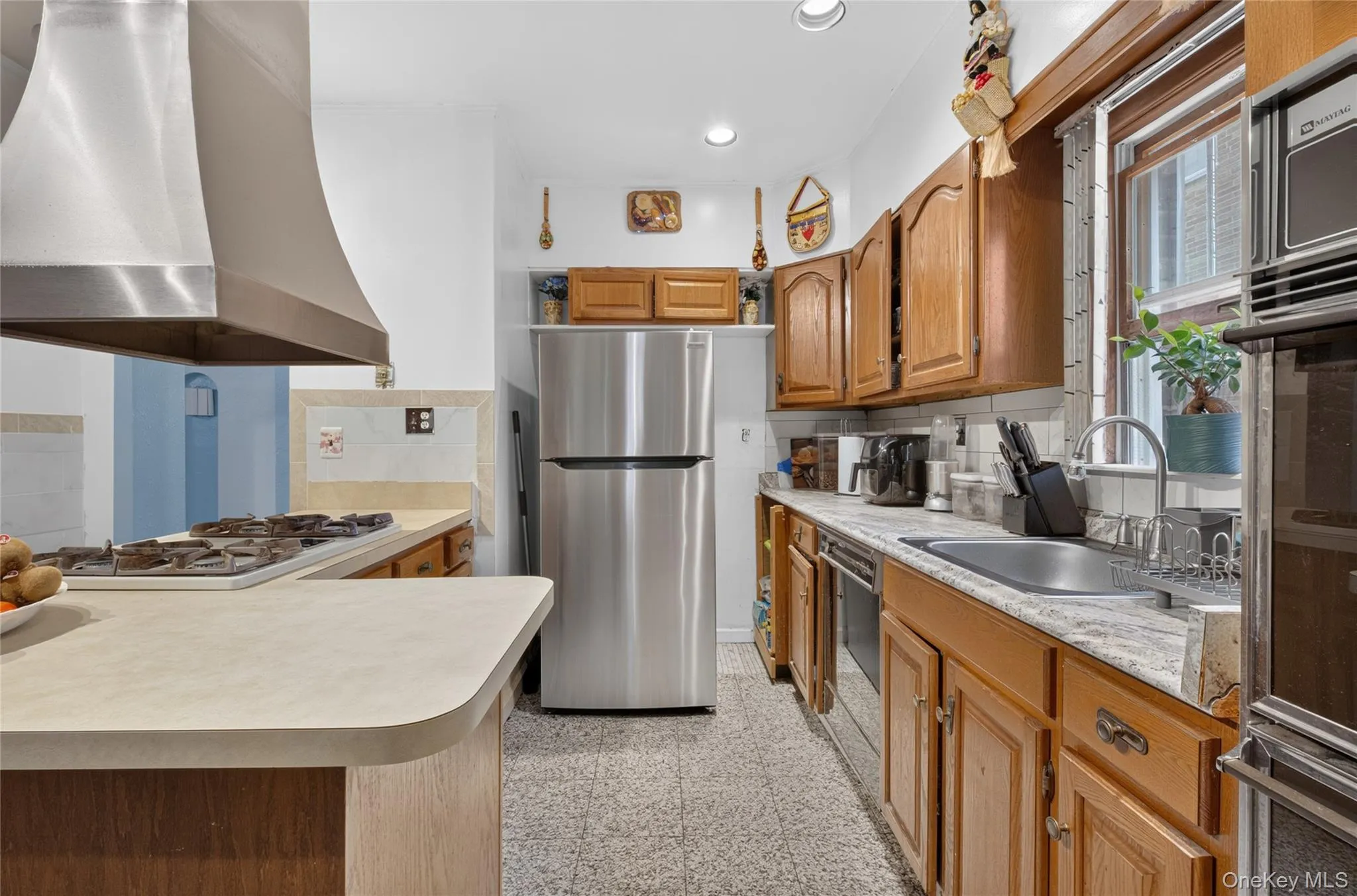 Galley kitchen featuring wood cabinetry, stainless steel refrigerator, stainless steel range hood, built-in stovetop, and built-in oven Galley kitchen featuring wood cabinetry, stainless steel refrigerator, stainless steel range hood, built-in stovetop, and built-in oven