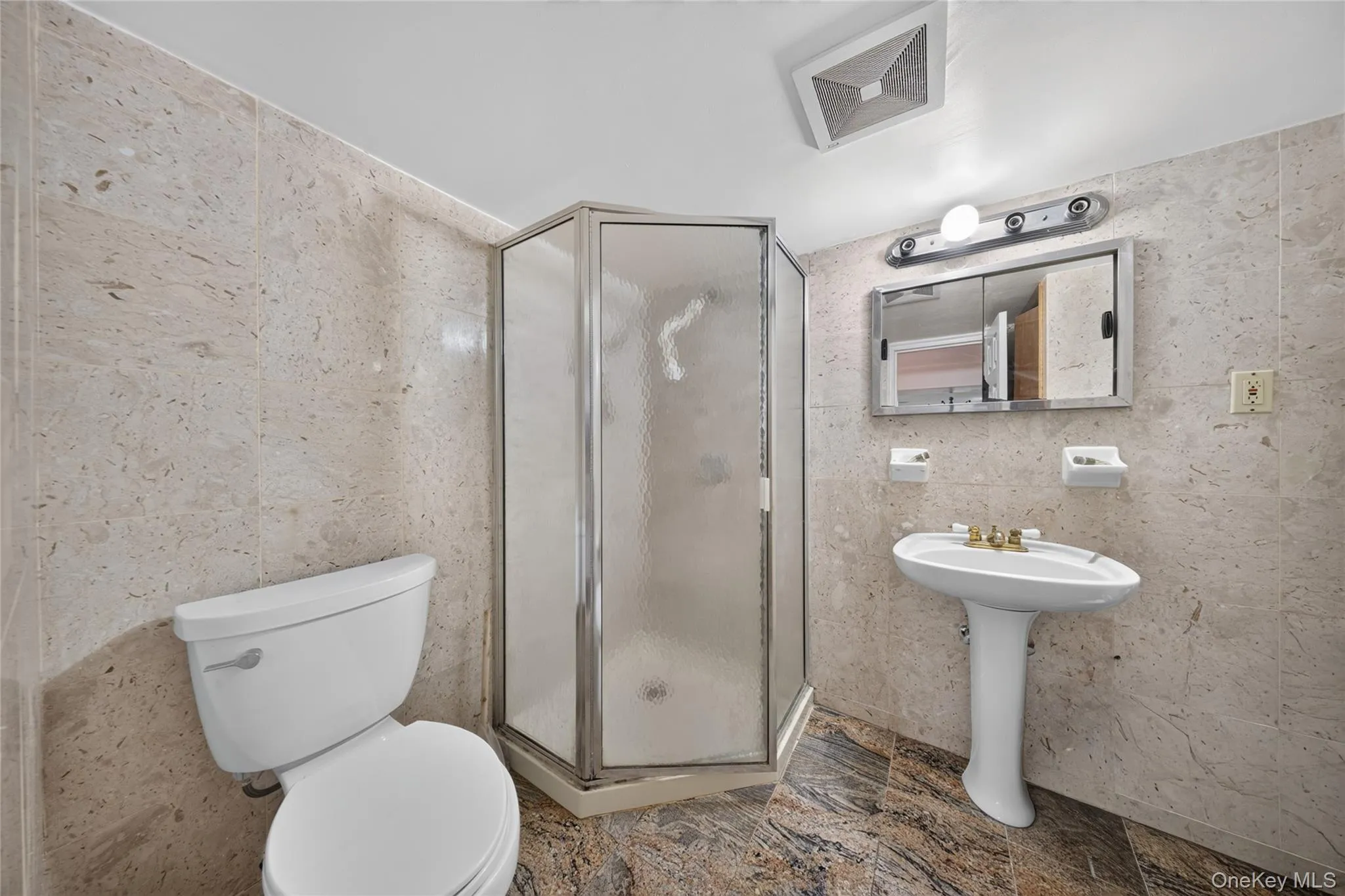 Bathroom featuring floor-to-ceiling stone-look tile, corner shower enclosure with frosted glass, white pedestal sink, and a white toilet Bathroom featuring floor-to-ceiling stone-look tile, corner shower enclosure with frosted glass, white pedestal sink, and a white toilet